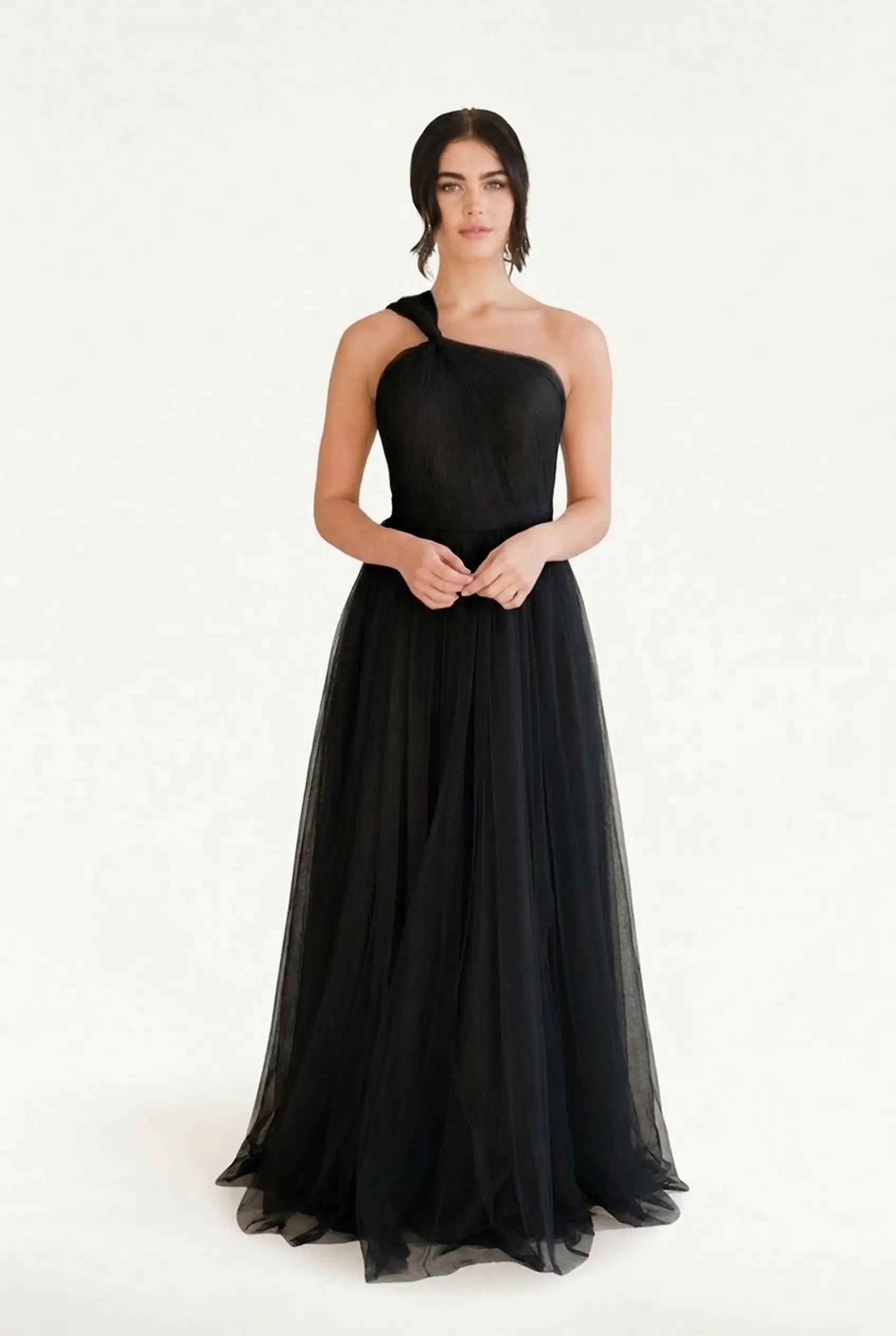 A woman poses against a white background in the TH&TH Luna Bridesmaid Dress in Black Tulle, featuring a long one-shoulder design with a flowing tulle skirt—an elegant, modern choice for bridal parties.