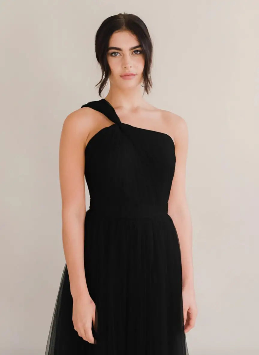 A woman models the Luna Bridesmaid Dress in Black Tulle by TH&TH against a neutral background. Her dark hair is styled in a loose updo, highlighting the elegant simplicity ideal for modern bridal parties.