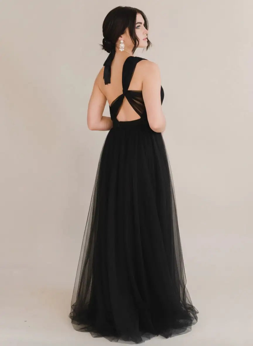 A woman wears the TH&TH Luna Bridesmaid Dress in Black Tulle, featuring crisscrossed straps and an open back—ideal for modern bridal parties. Her hair is in a low ribbon updo with statement earrings, set against a plain light background.