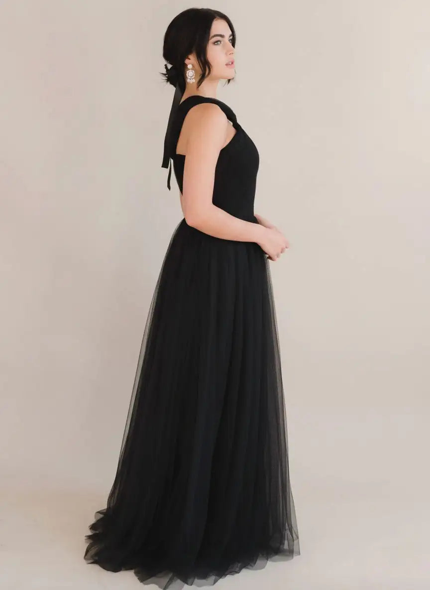 A woman in profile wears the Luna Bridesmaid Dress in Black Tulle by TH&TH—sleeveless with a floor-length skirt. Her updo, black ribbon, and statement earrings complete this look, ideal for modern bridal parties against a plain light backdrop.