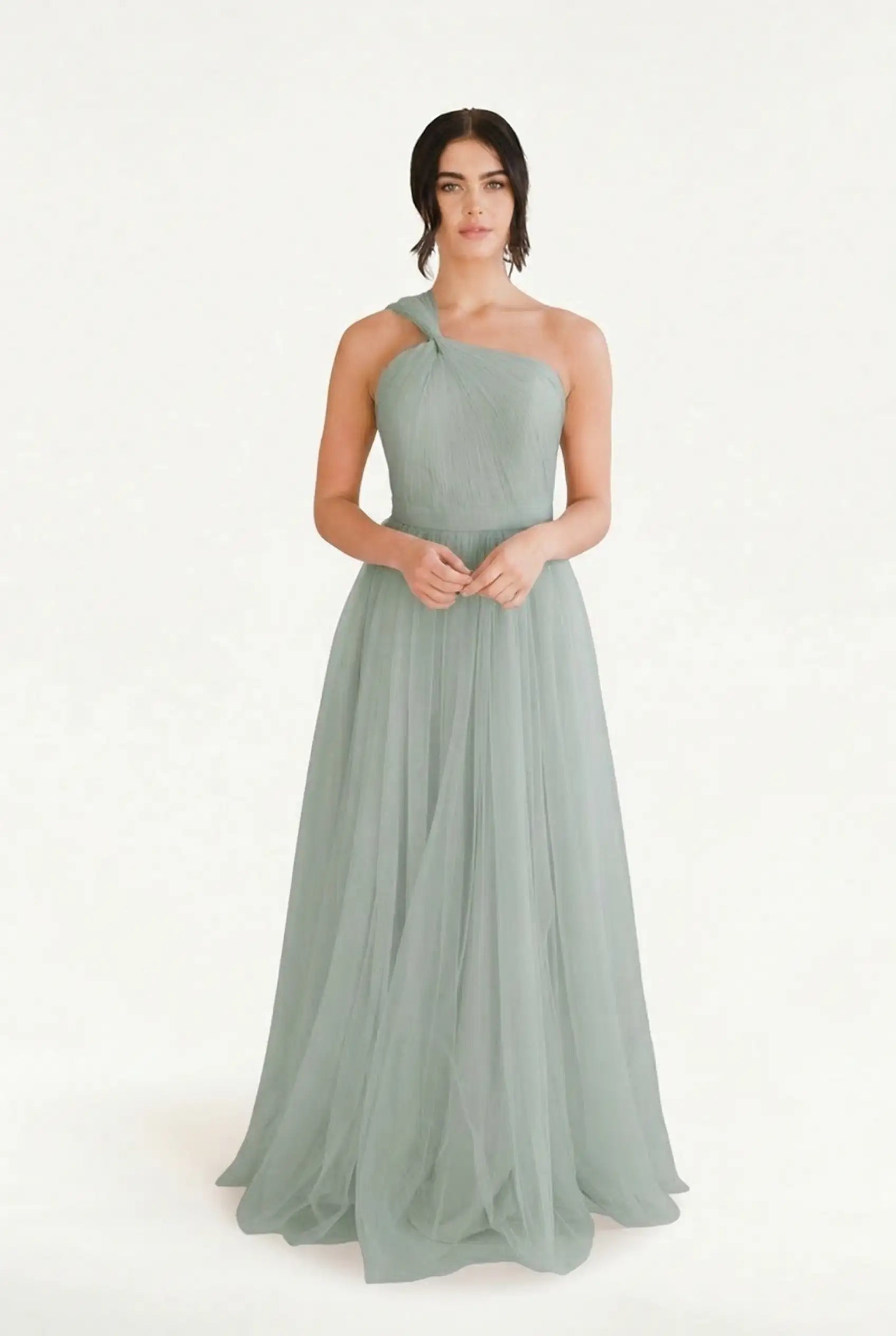 A woman stands against a plain background wearing the TH&TH Luna Bridesmaid Dress in Dusty Eucalyptus Tulle, a long, one-shoulder gown with gathered details—an elegant modern choice. Her hair is tied back as she looks straight ahead.