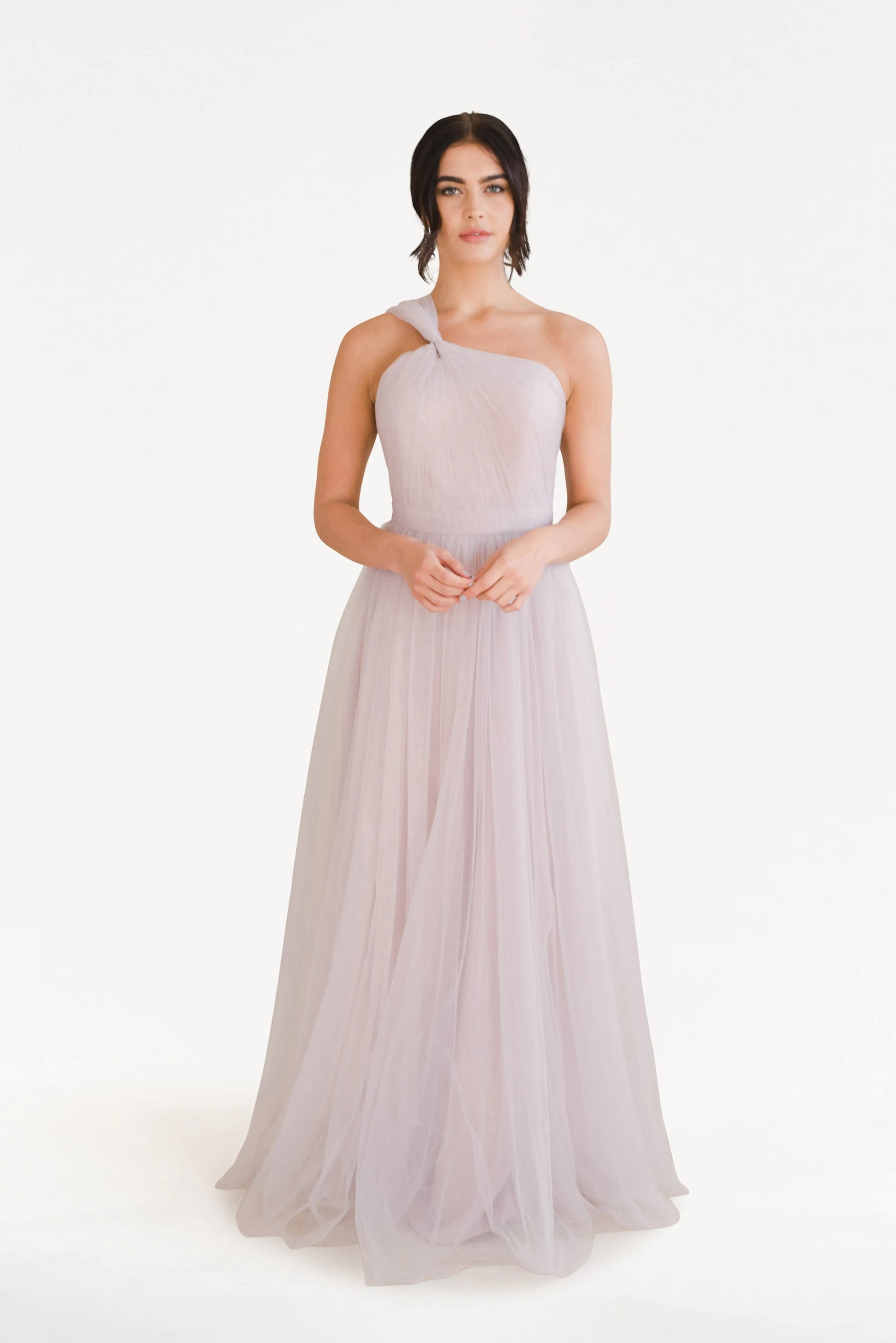 A woman with dark hair styled back stands against a white background, hands clasped, wearing the TH&TH Luna Bridesmaid Dress in Smoked Blush Tulle—a long, flowing, one-shoulder gown crafted from sheer tulle.