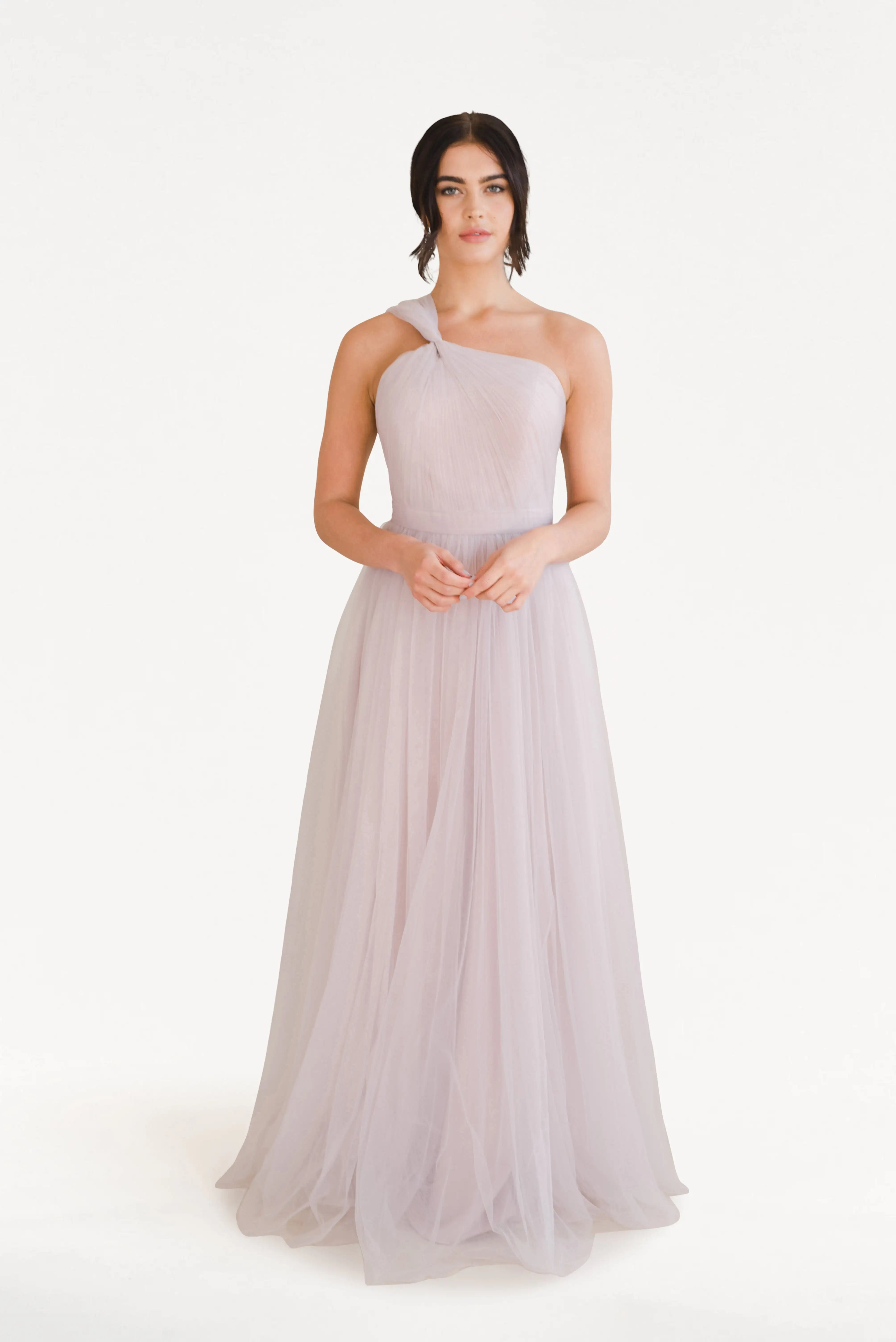 A woman with dark hair styled back stands against a white background, hands clasped, wearing the TH&TH Luna Bridesmaid Dress in Smoked Blush Tulle—a long, flowing, one-shoulder gown crafted from sheer tulle.