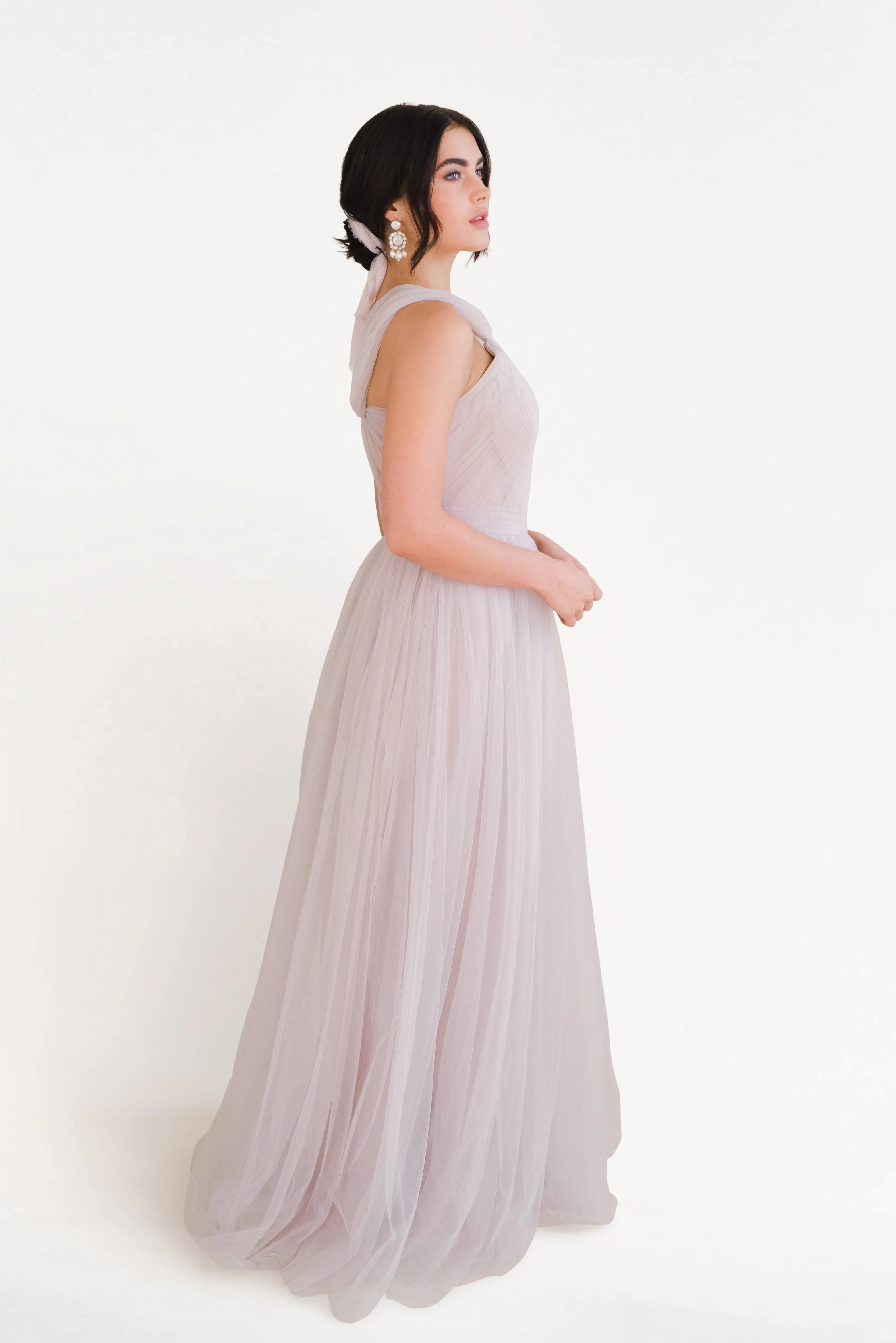 A woman stands in profile against a white background wearing the TH&TH Luna Bridesmaid Dress in Smoked Blush Tulle, with her dark hair styled up with a ribbon and statement earrings.