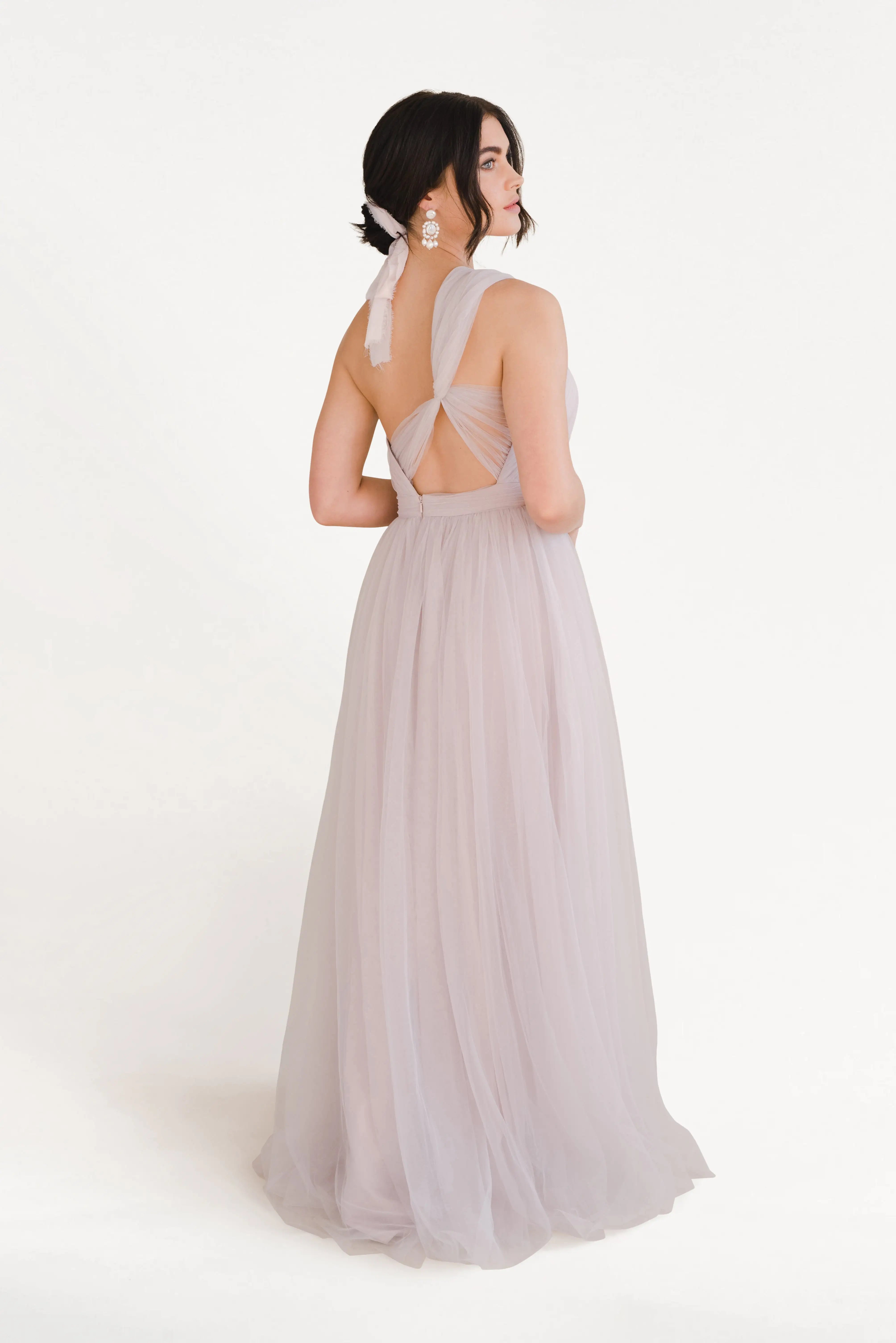 A woman with dark hair in a low bun models the TH&TH Luna Bridesmaid Dress in smoked blush tulle, featuring crisscross straps and an open back, standing against a plain white background.