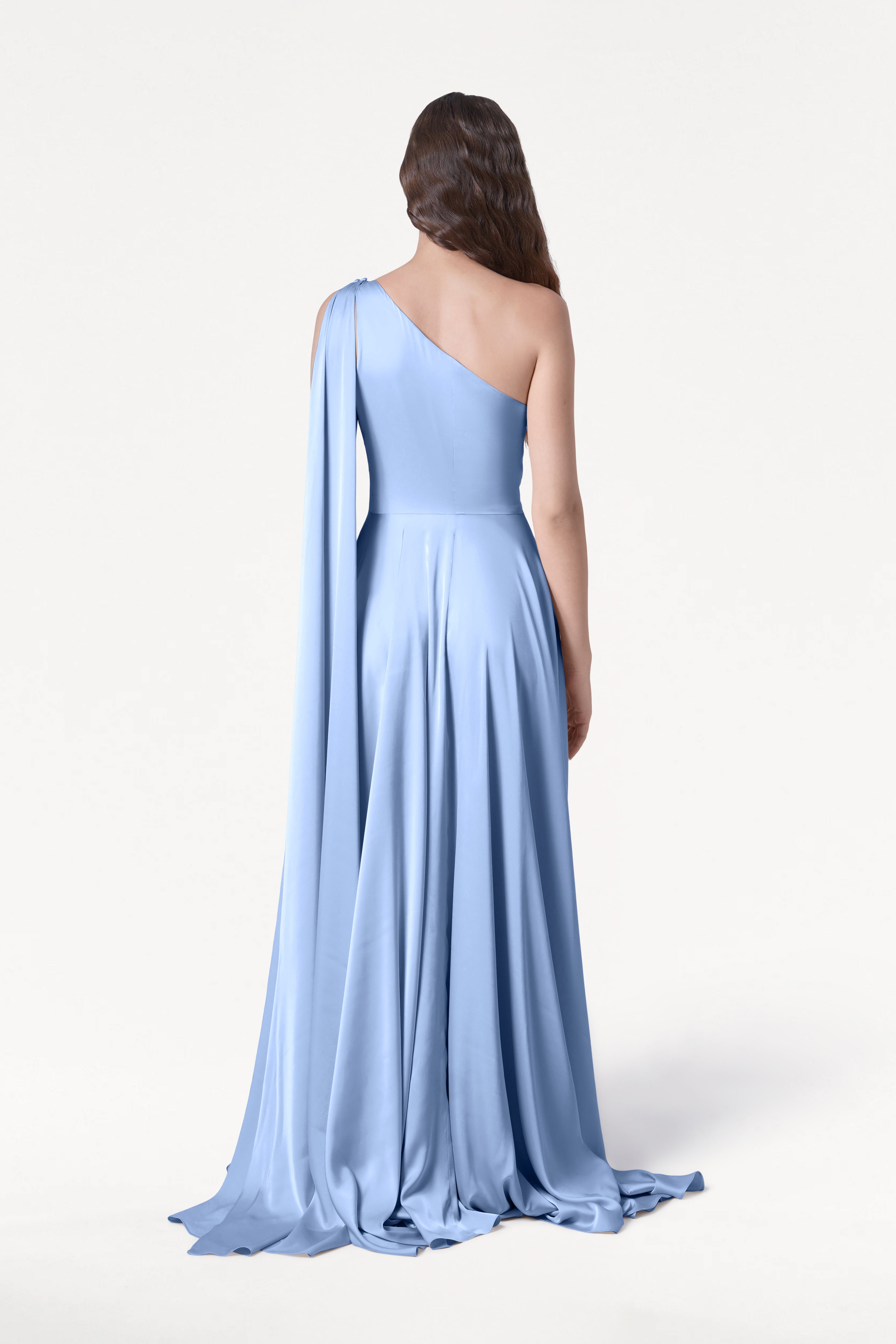A woman with loose waves faces away in the Lyra Bridesmaid Dress by TH&TH, featuring light blue satin, one-shoulder draped sleeve, and a size-inclusive fit. The background is plain white, highlighting the gown's effortless elegance.