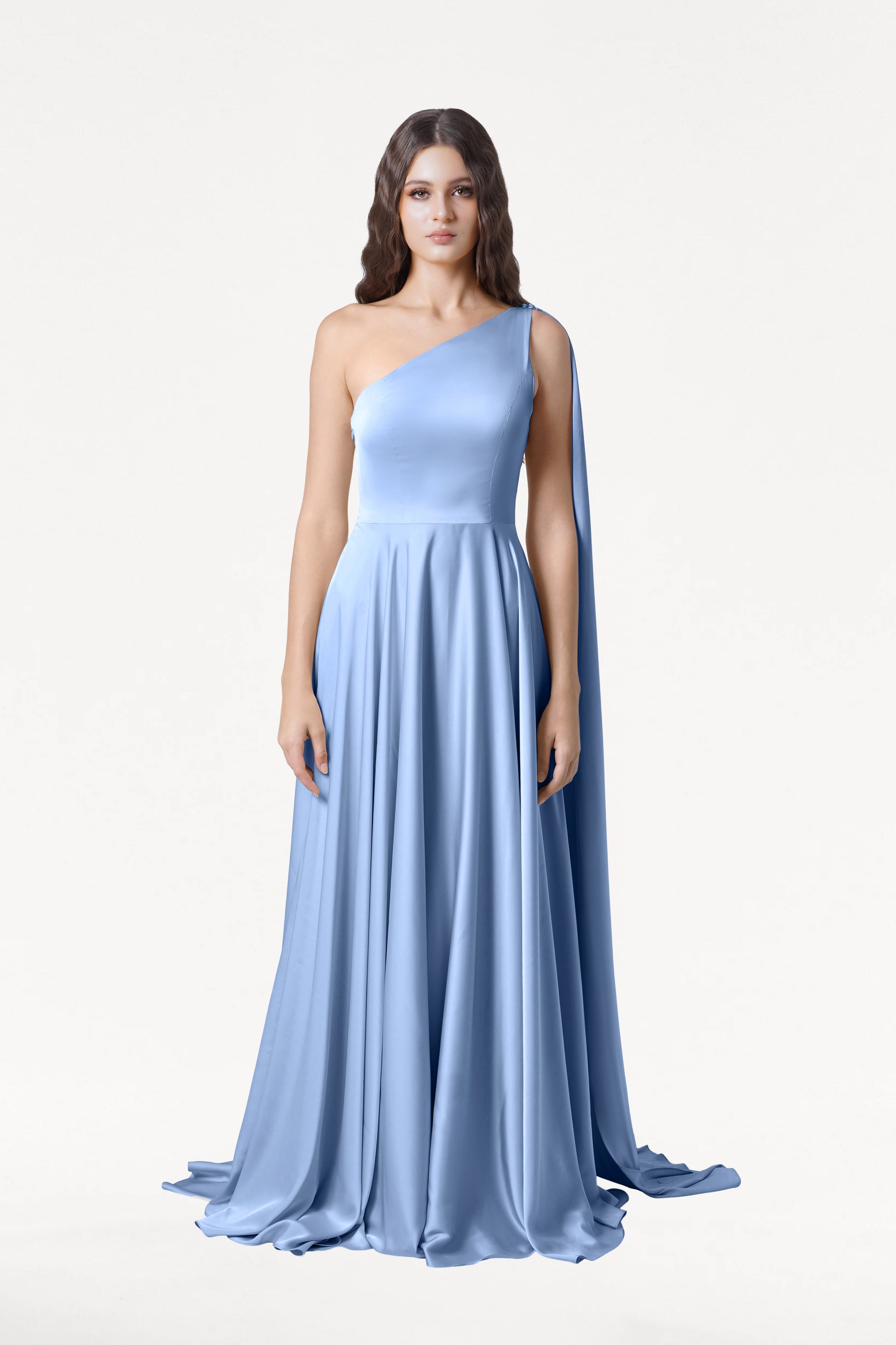 A woman wears the Lyra Bridesmaid Dress in Dusty Blue Satin by TH&TH, featuring a long, one-shoulder design with a flowing skirt and draped cape, set against a plain white background.