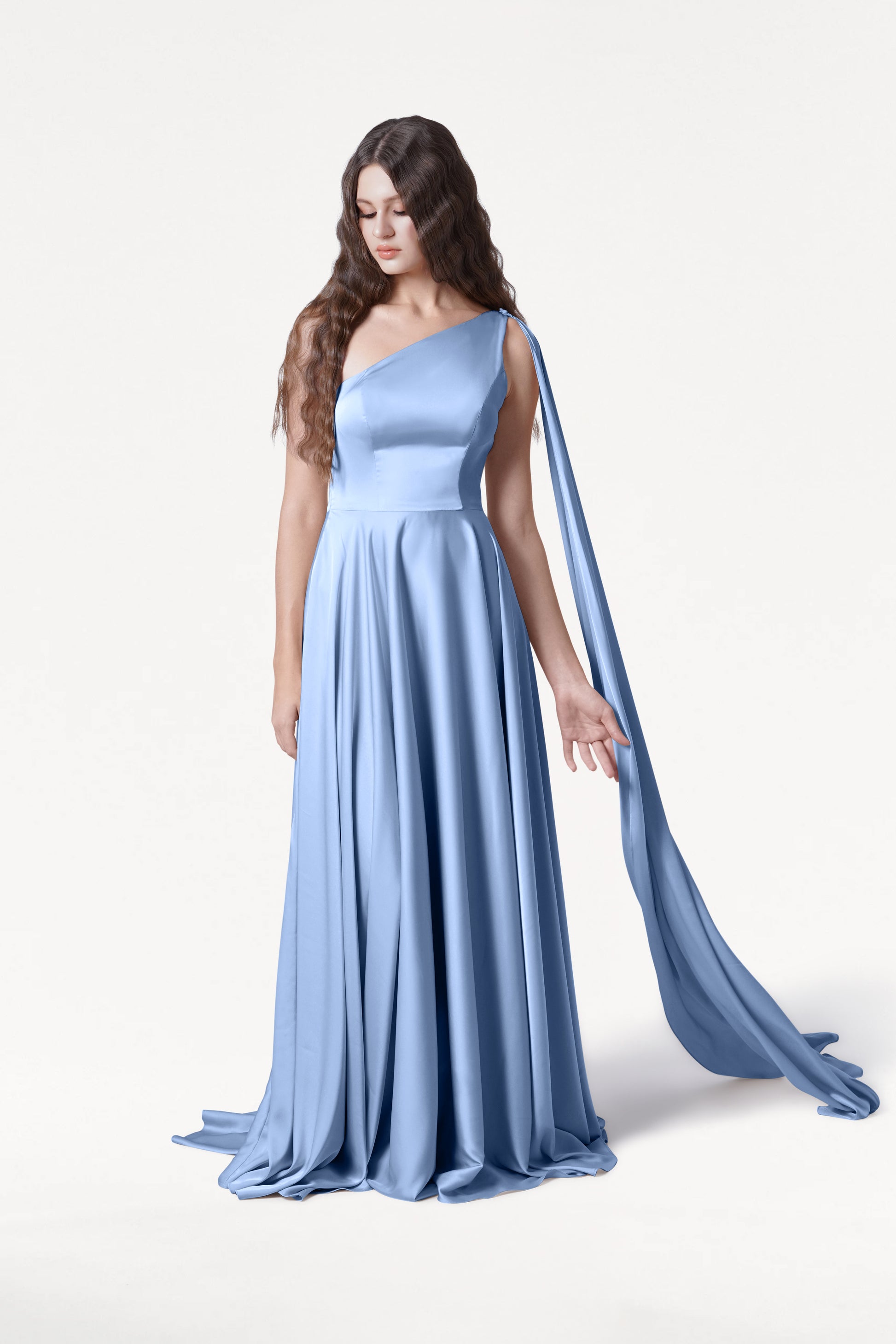 A woman with long, wavy brown hair models the Lyra Bridesmaid Dress in Dusty Blue Satin by TH&TH, featuring a flowing one-shoulder design with a draped sash. She stands gracefully against a plain white background, gazing down and to the side.