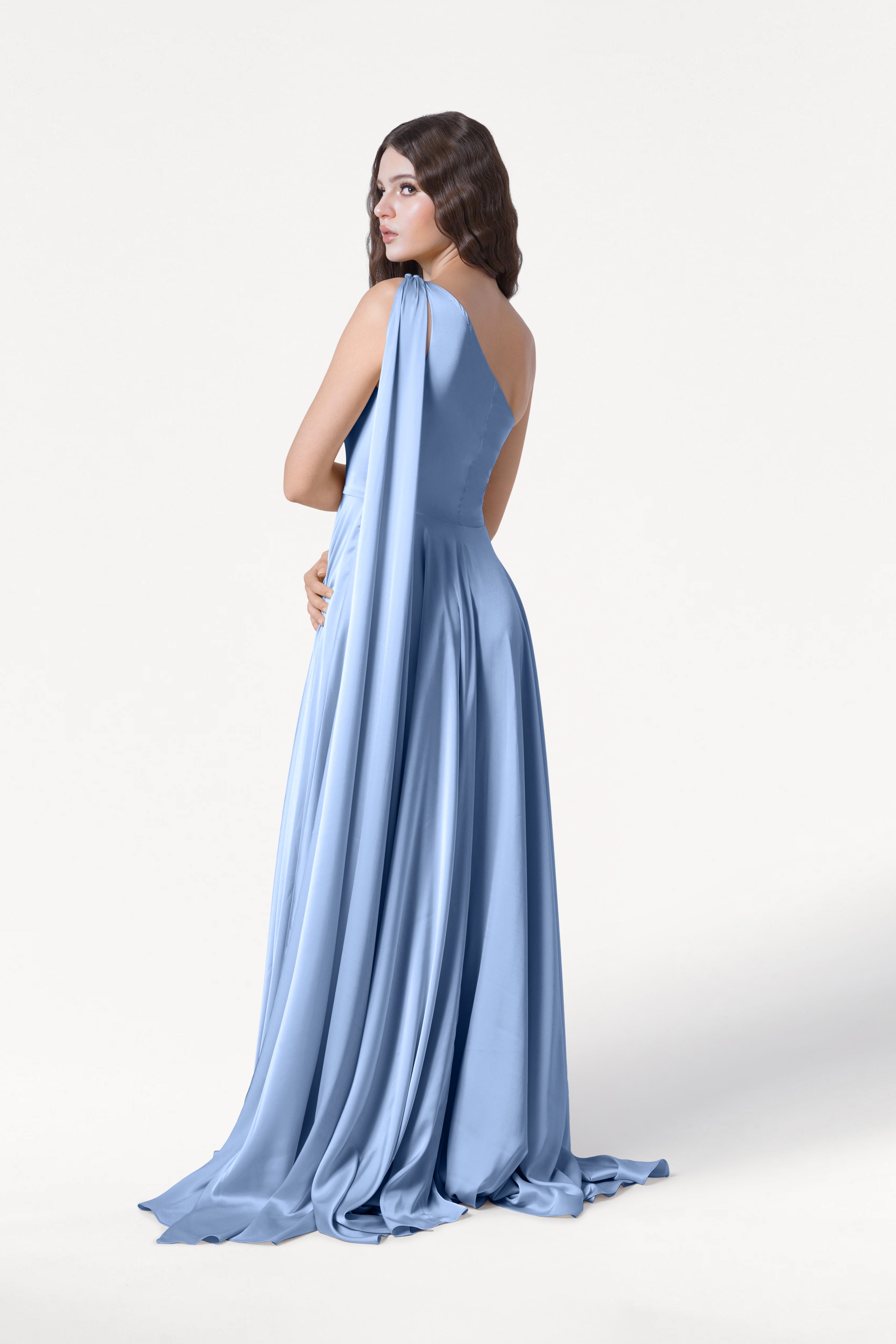 A woman models the TH&TH Lyra Bridesmaid Dress in Dusty Blue Satin, a modern, floor-length gown accentuating her figure as she stands against a white background, looking over her shoulder with one arm at her side.