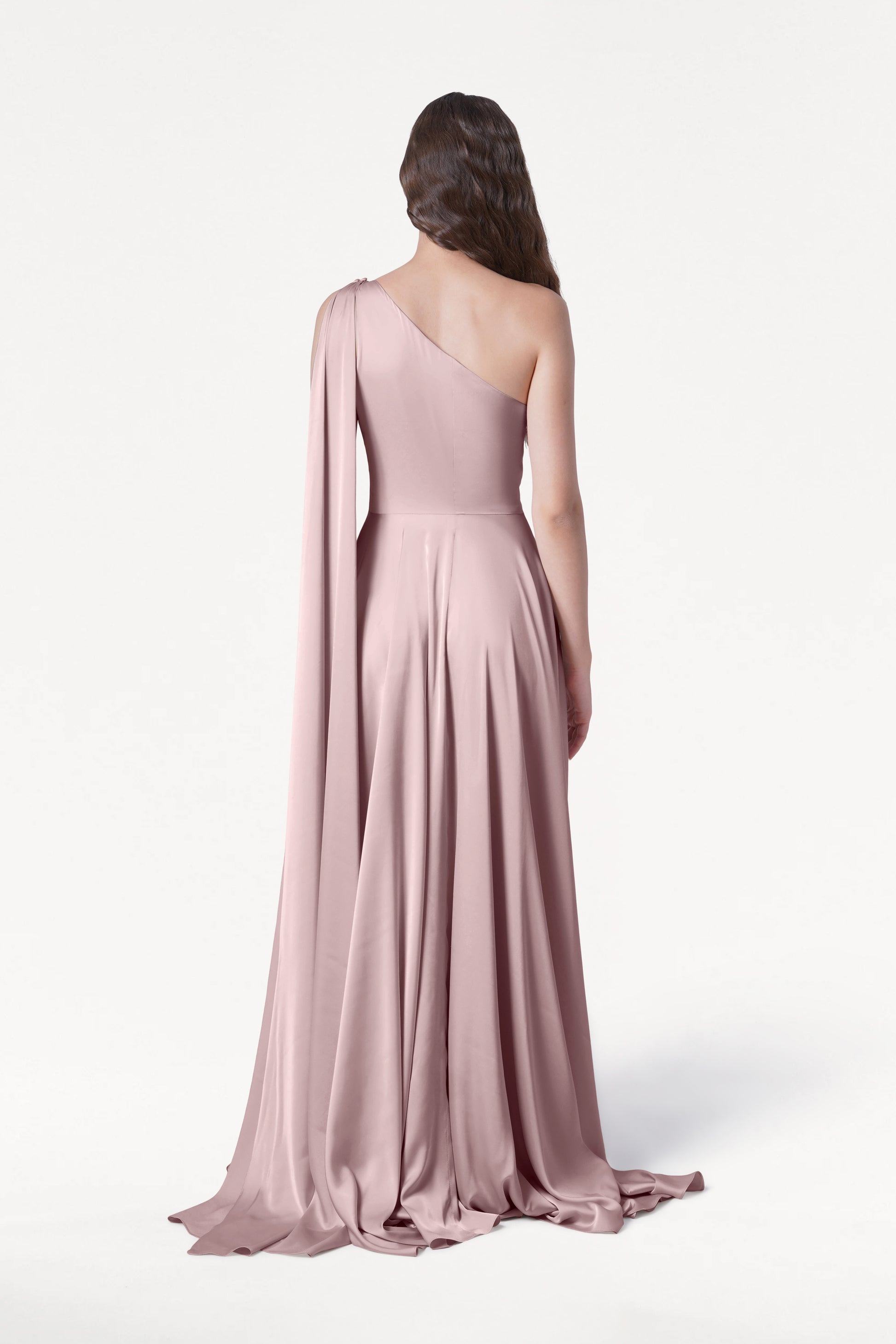 A person faces away, wearing the size-inclusive TH&TH Lyra Bridesmaid Dress in Smoked Blush Satin—a floor-length, one-shoulder gown with a flowing drape—exuding smooth elegance against a plain white background.