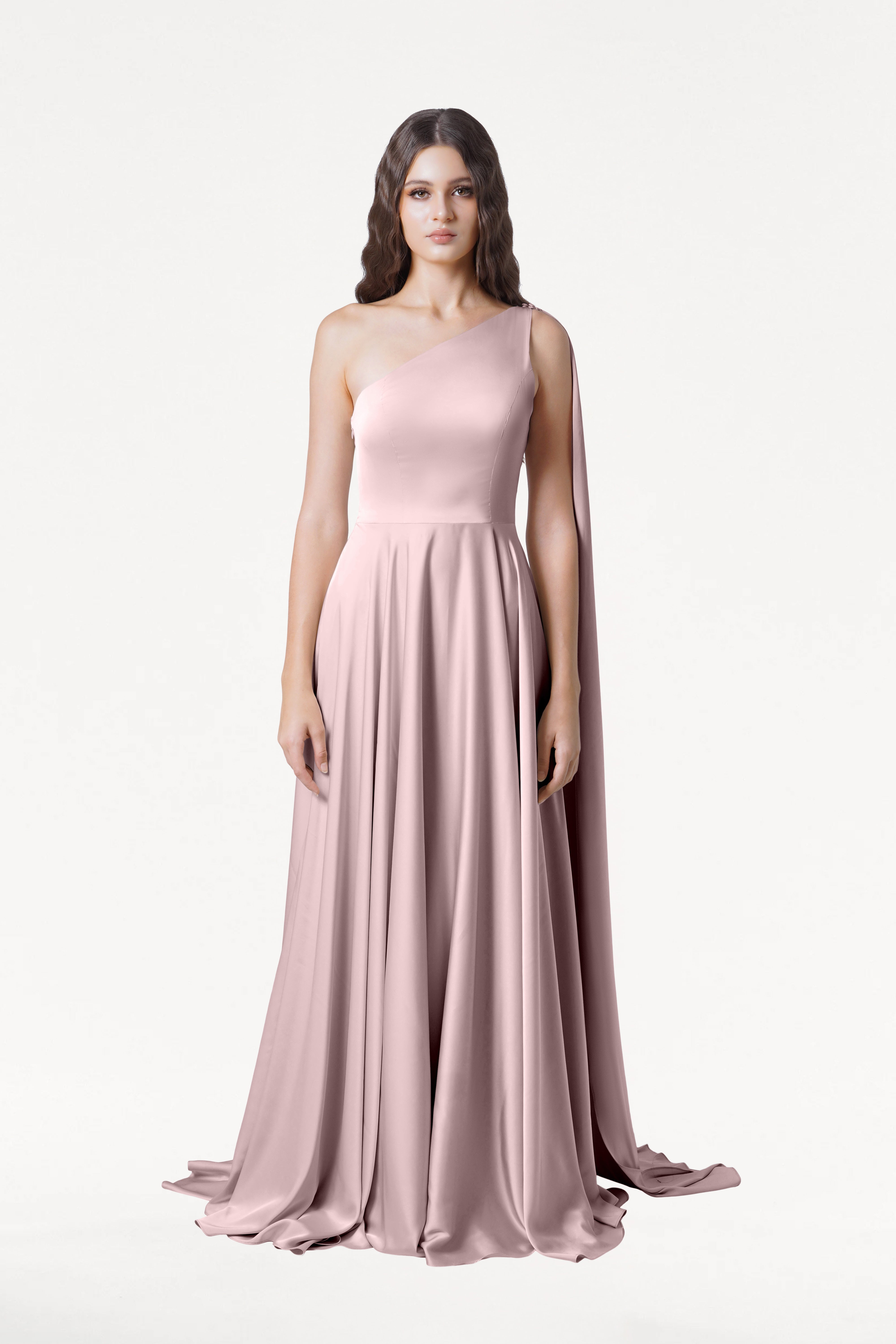 A woman stands against a plain white background in the TH&TH Lyra Bridesmaid Dress in Smoked Blush Satin, a size-inclusive, floor-length one-shoulder gown with a flowing skirt and draped fabric over one arm.