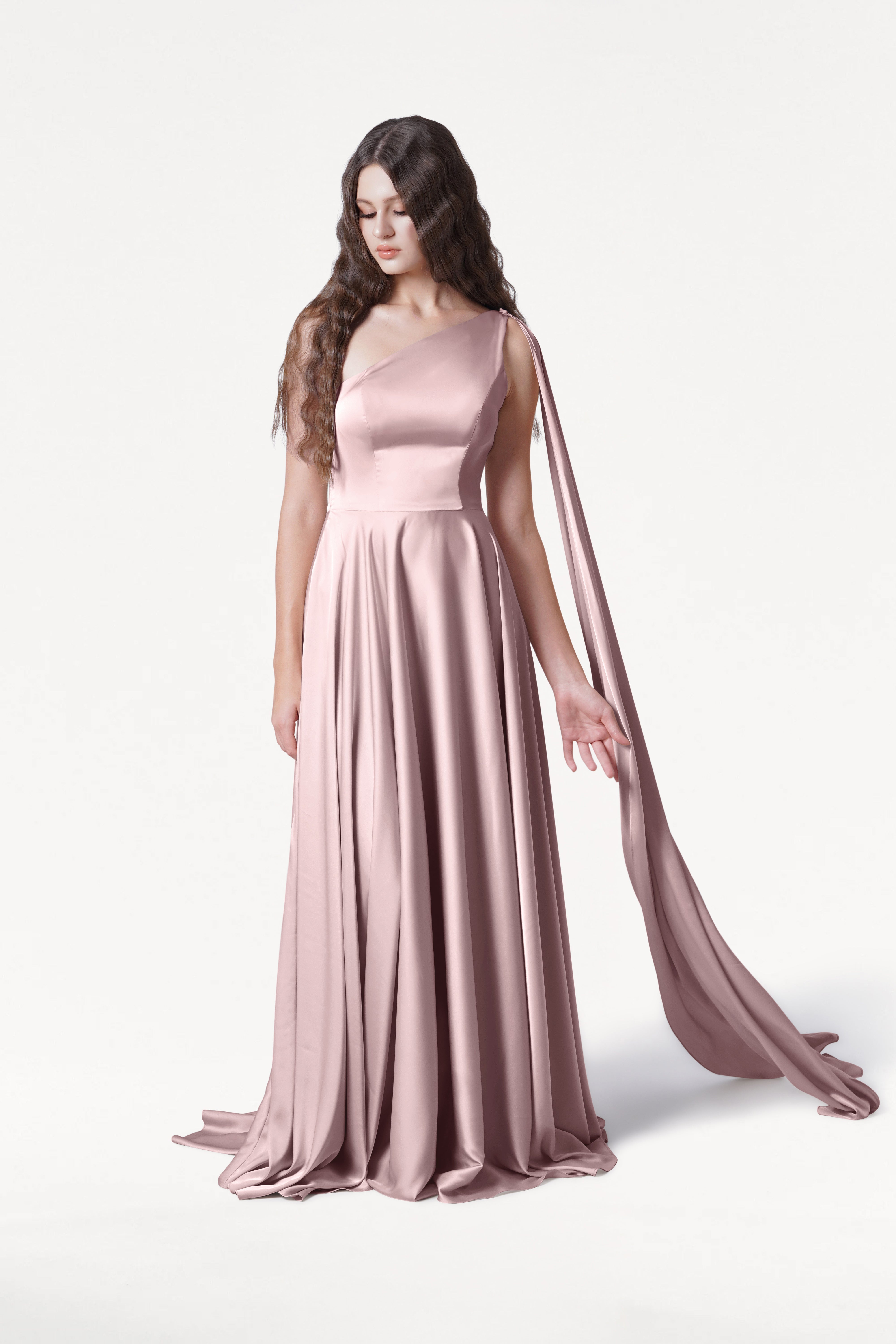 A woman with long wavy brown hair wears the TH&TH Lyra Bridesmaid Dress in Smoked Blush Satin, featuring a single shoulder strap and long draped fabric, standing against a plain white background.
