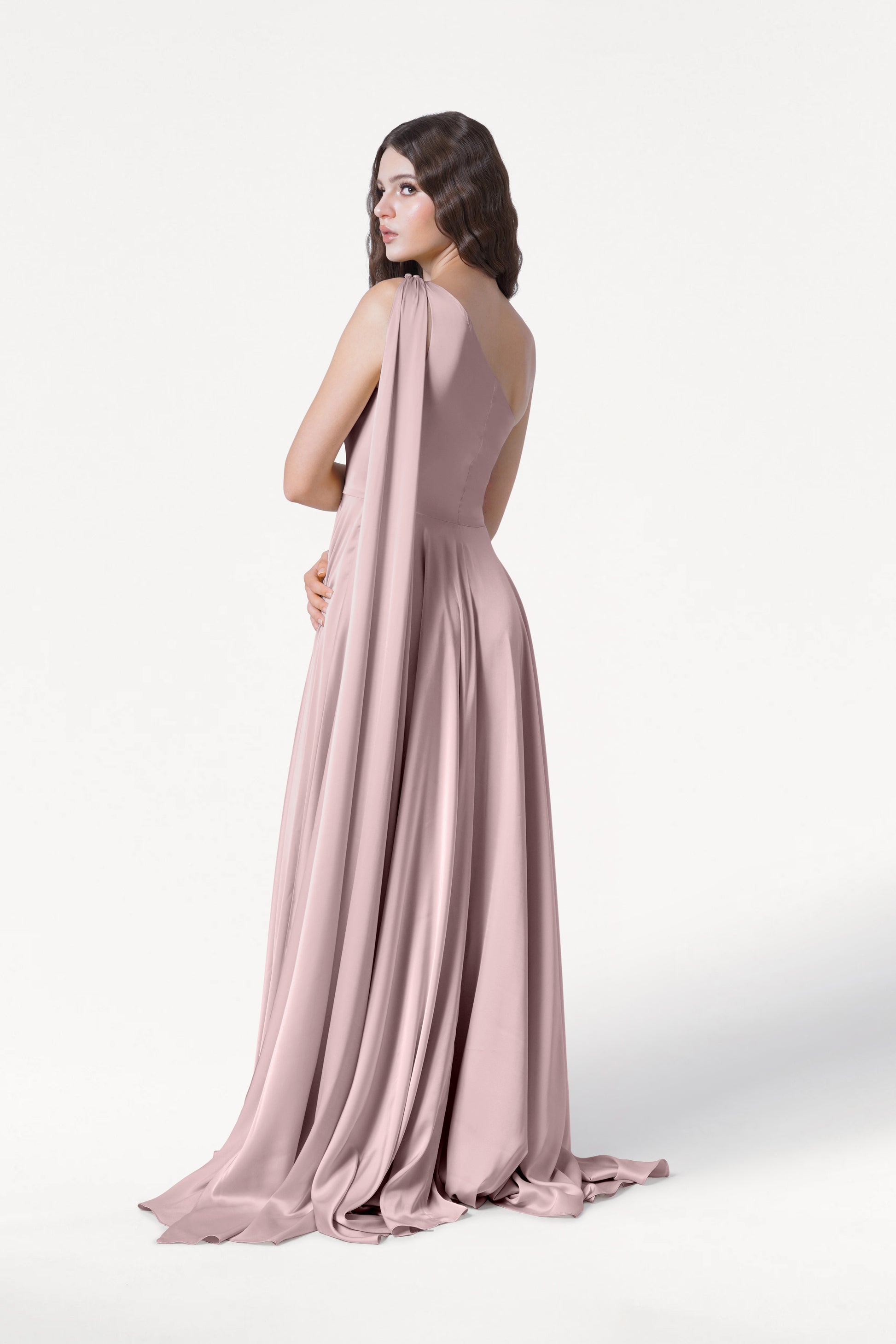 A woman with long wavy brown hair models the TH&TH Lyra Bridesmaid Dress in Smoked Blush Satin—a size-inclusive, floor-length gown with a flowing train and draped fabric—standing against a plain white background, looking over her shoulder.