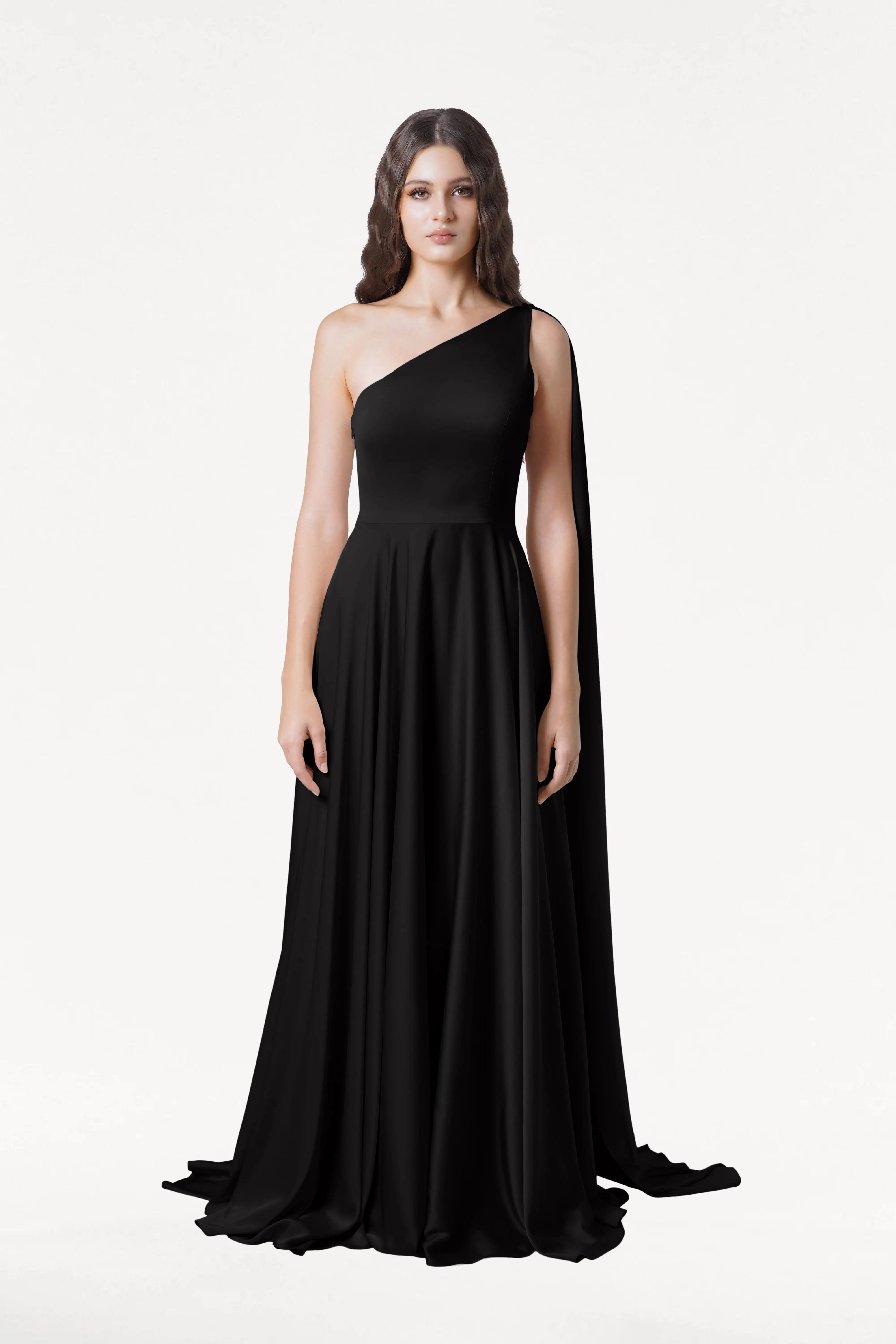 A woman with long wavy brown hair models the TH&TH Lyra Bridesmaid Dress in Black Satin—a floor-length, one-shoulder gown with a flowing skirt—posed against a plain white background for an effortlessly elegant look.