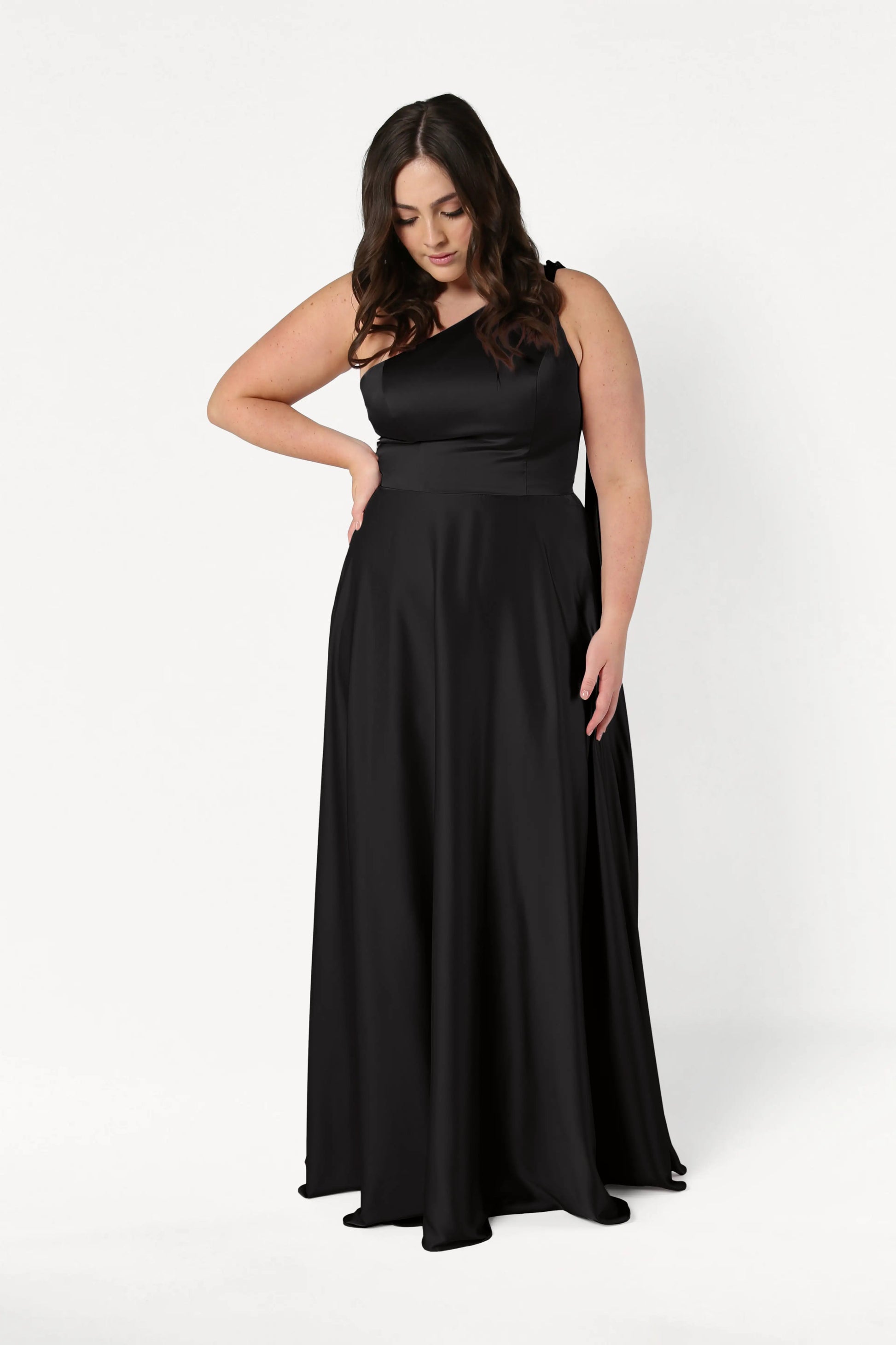 A woman with long dark hair wears the TH&TH Lyra Bridesmaid Dress in Black Satin, a floor-length, one-shoulder style. She stands against a plain white background, looking down with one hand on her waist.