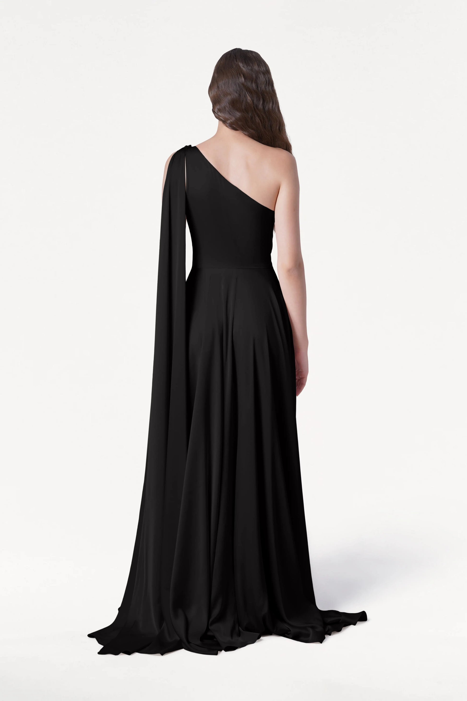 A woman faces away, wearing the Lyra Bridesmaid Dress in Black Satin by TH&TH—a long, flowing one-shoulder gown with a draped sleeve. Her wavy hair falls down her back against a plain white background.
