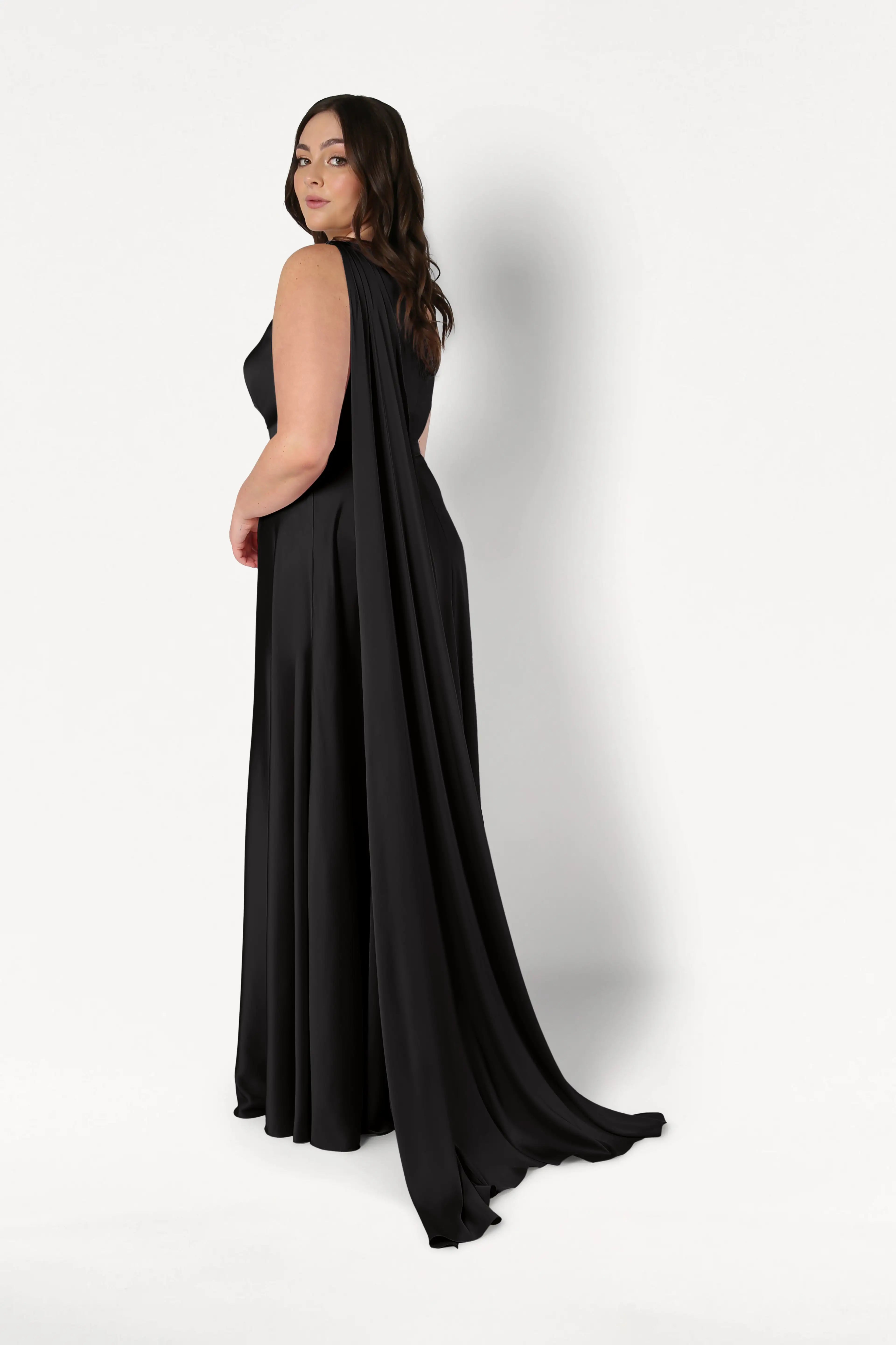 A woman models the TH&TH Lyra Bridesmaid Dress in Black Satin, a sleeveless, floor-length gown with a long draped cape, as she stands against a white background and looks over her shoulder at the camera.