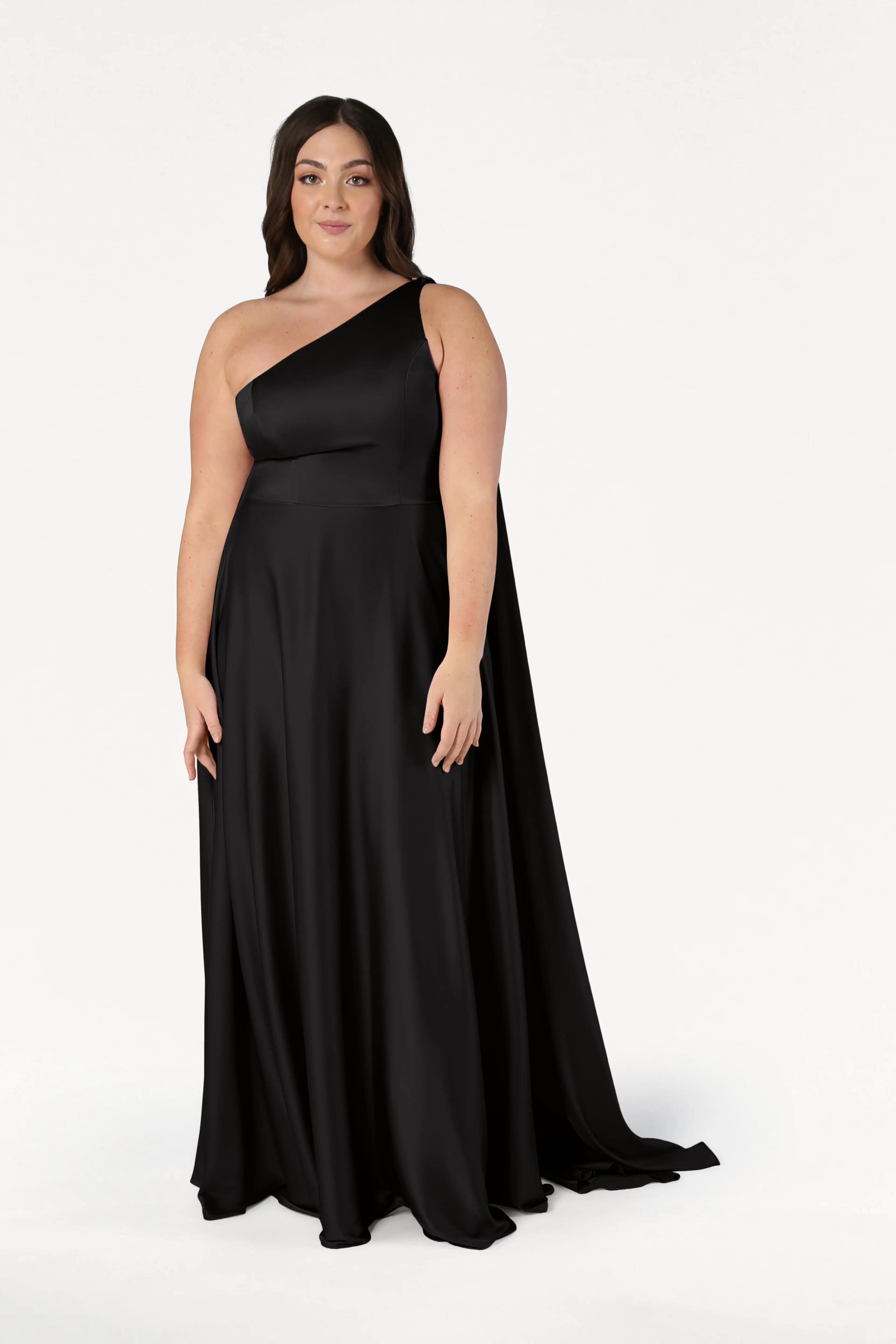 A woman with long dark hair and a neutral expression models the TH&TH Lyra Bridesmaid Dress in Black Satin, featuring a one-shoulder design and flowing skirt, posed against a plain white background.
