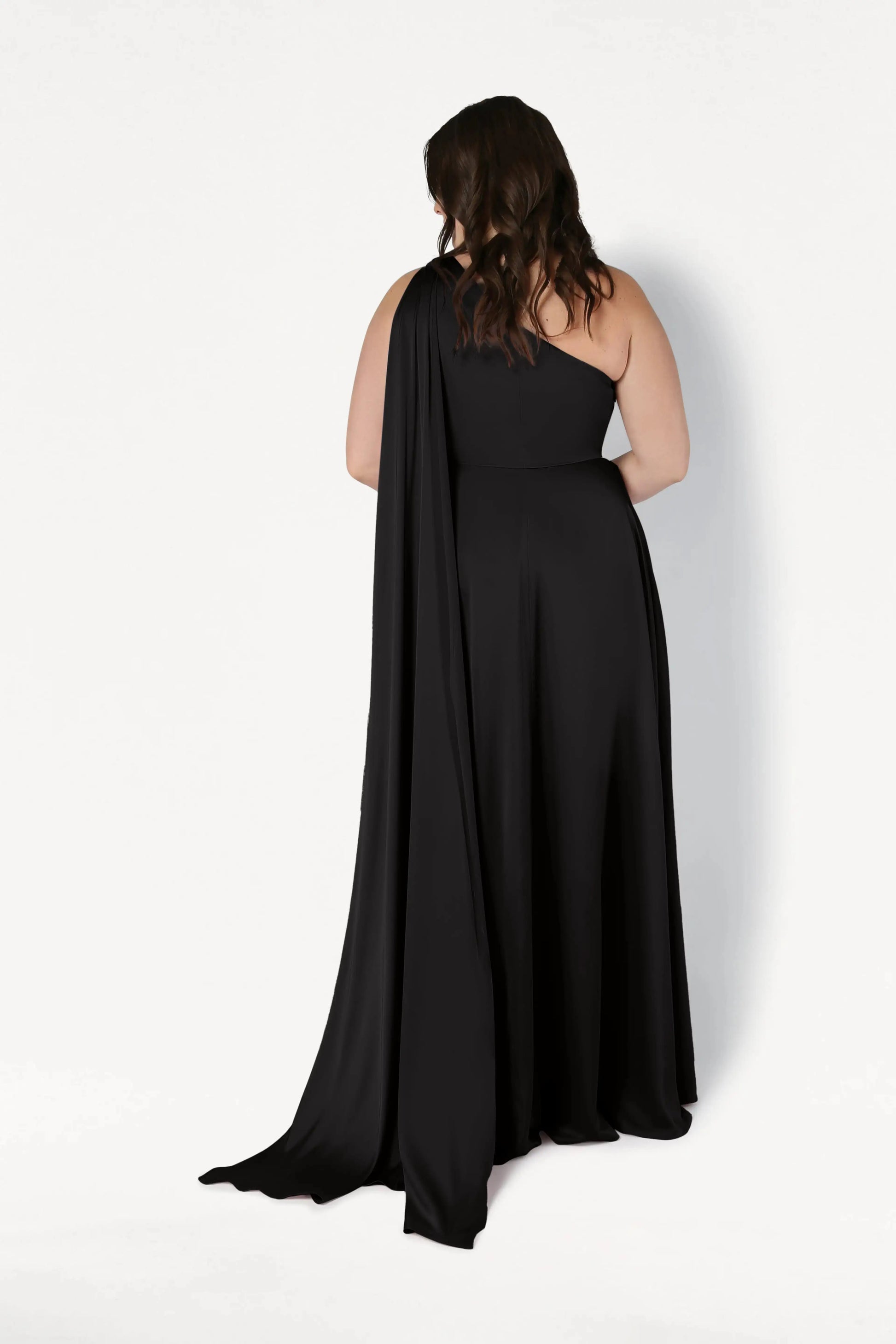 A woman with wavy brown hair stands facing away, wearing the TH&TH Lyra Bridesmaid Dress in black satin, featuring a modern one-shoulder design and draped cape, set against a plain white background.