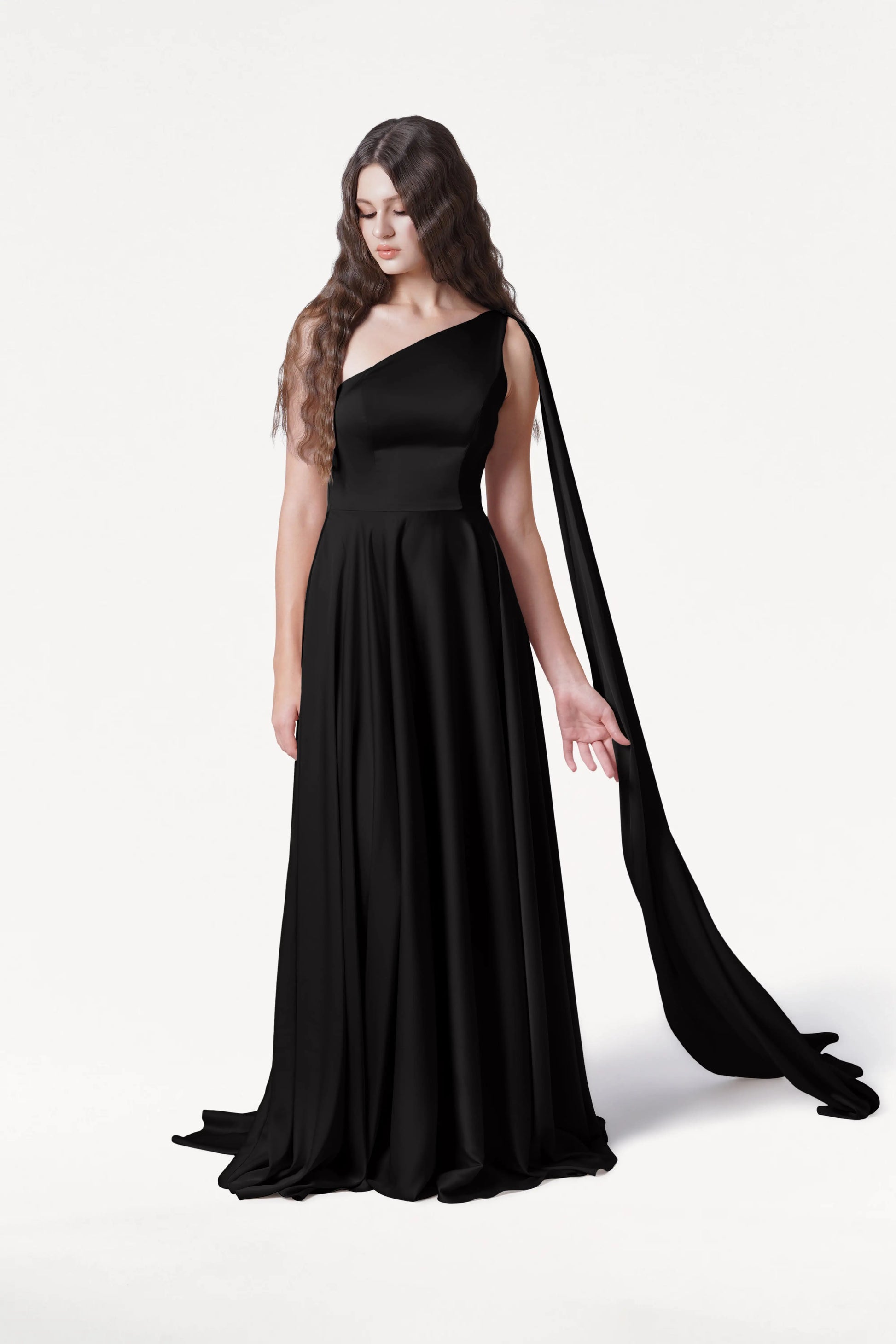 A woman with long wavy brown hair wears the Lyra Bridesmaid Dress in Black Satin by TH&TH, a size-inclusive one-shoulder gown with a draped sash, standing against a plain white background and looking slightly downward.