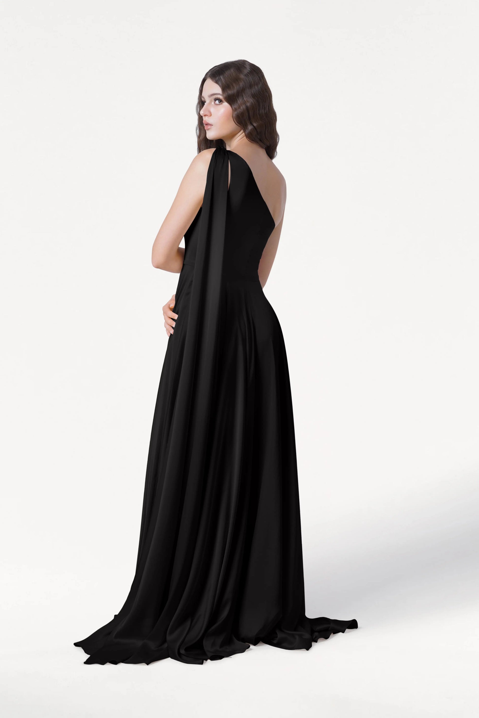 A woman with wavy brown hair models the Lyra Bridesmaid Dress in Black Satin by TH&TH, featuring a draped shoulder, as she looks over her shoulder against a plain white background.