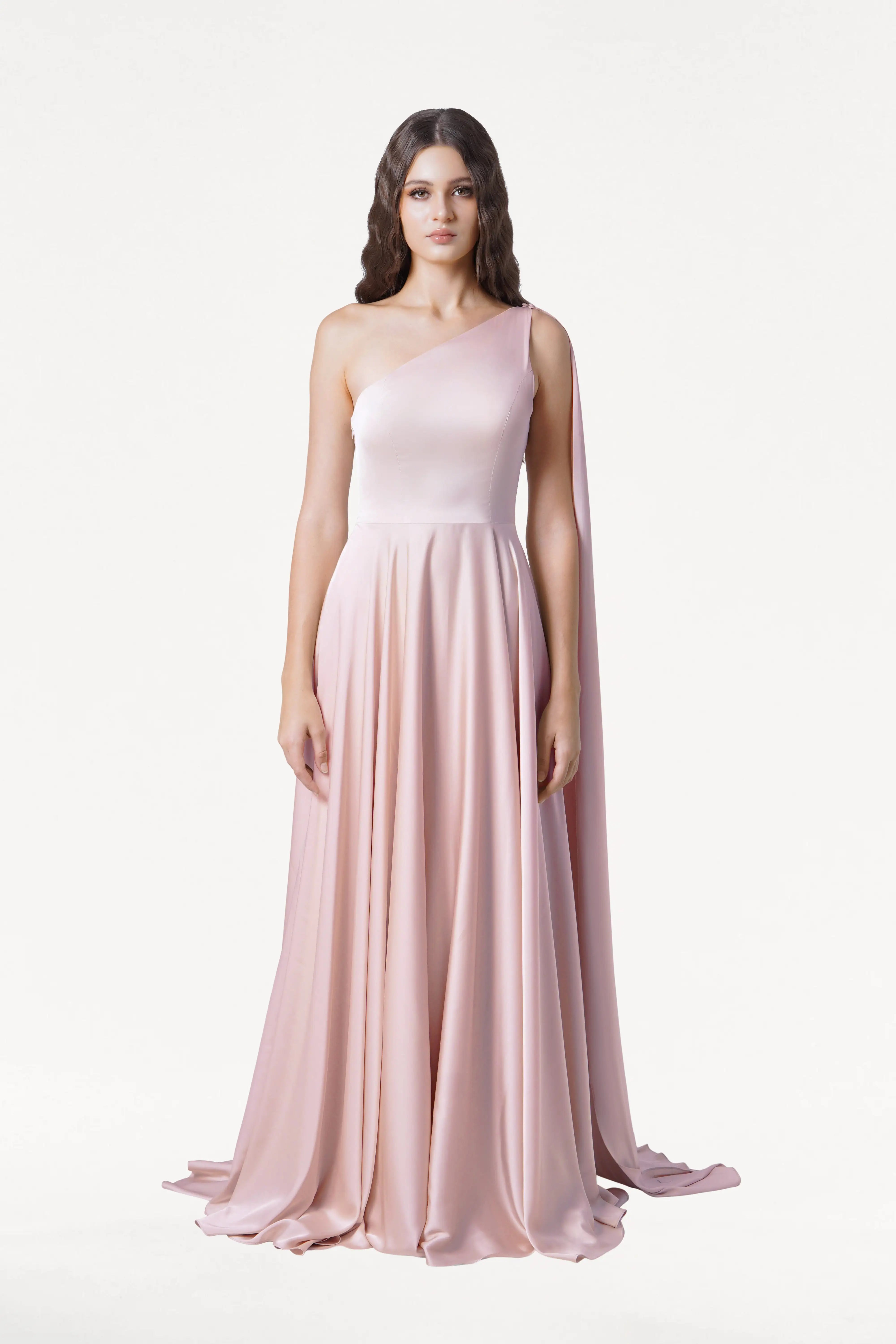 A woman with long wavy brown hair and a neutral expression models the TH&TH Lyra Bridesmaid Dress in Blush Pink Satin, a long one-shoulder gown with a flowing skirt and draped fabric, posing against a white background.