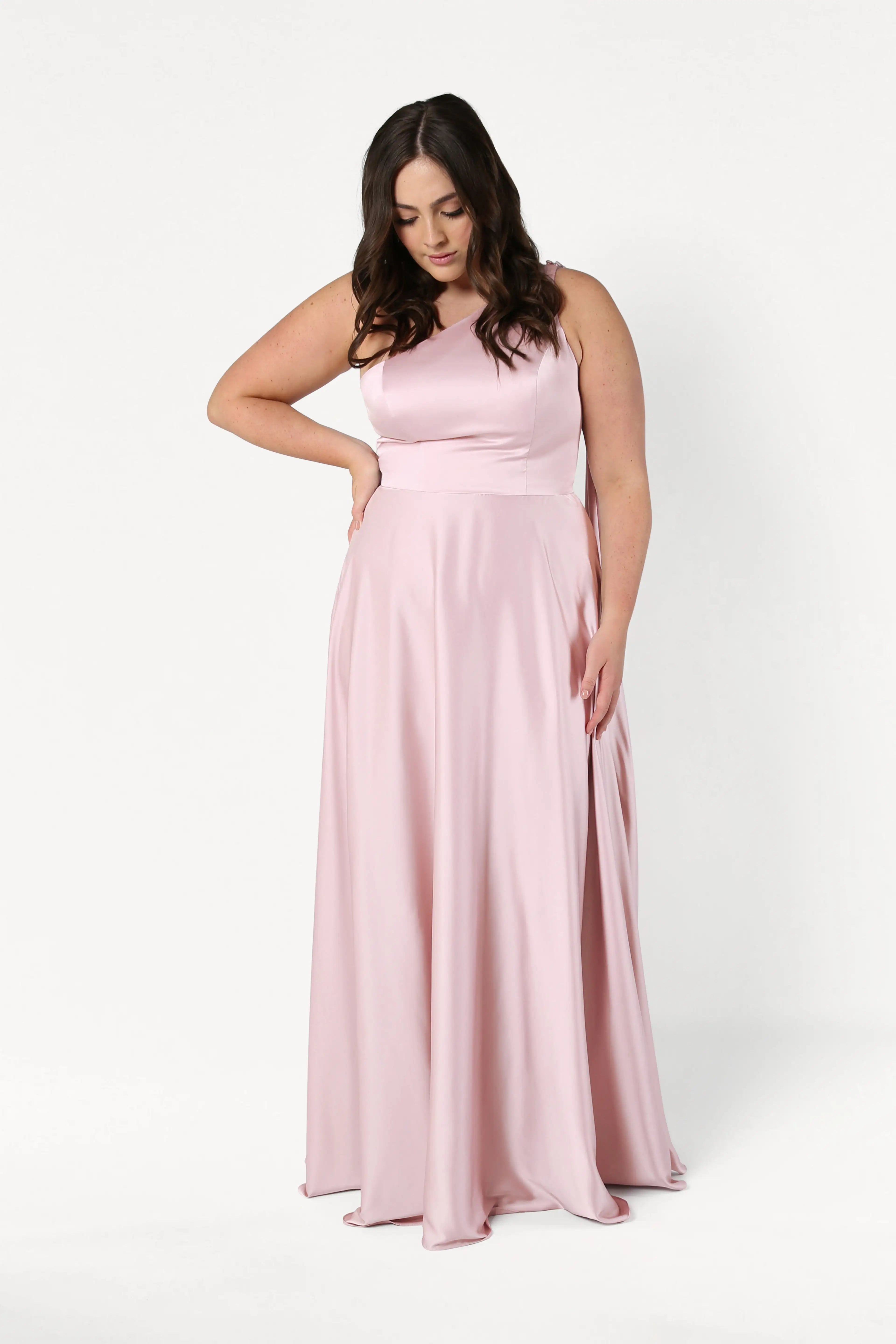 A woman wears the Lyra Bridesmaid Dress in Blush Pink Satin by TH&TH, standing against a white background with one hand on her hip and the other gently touching the dress.