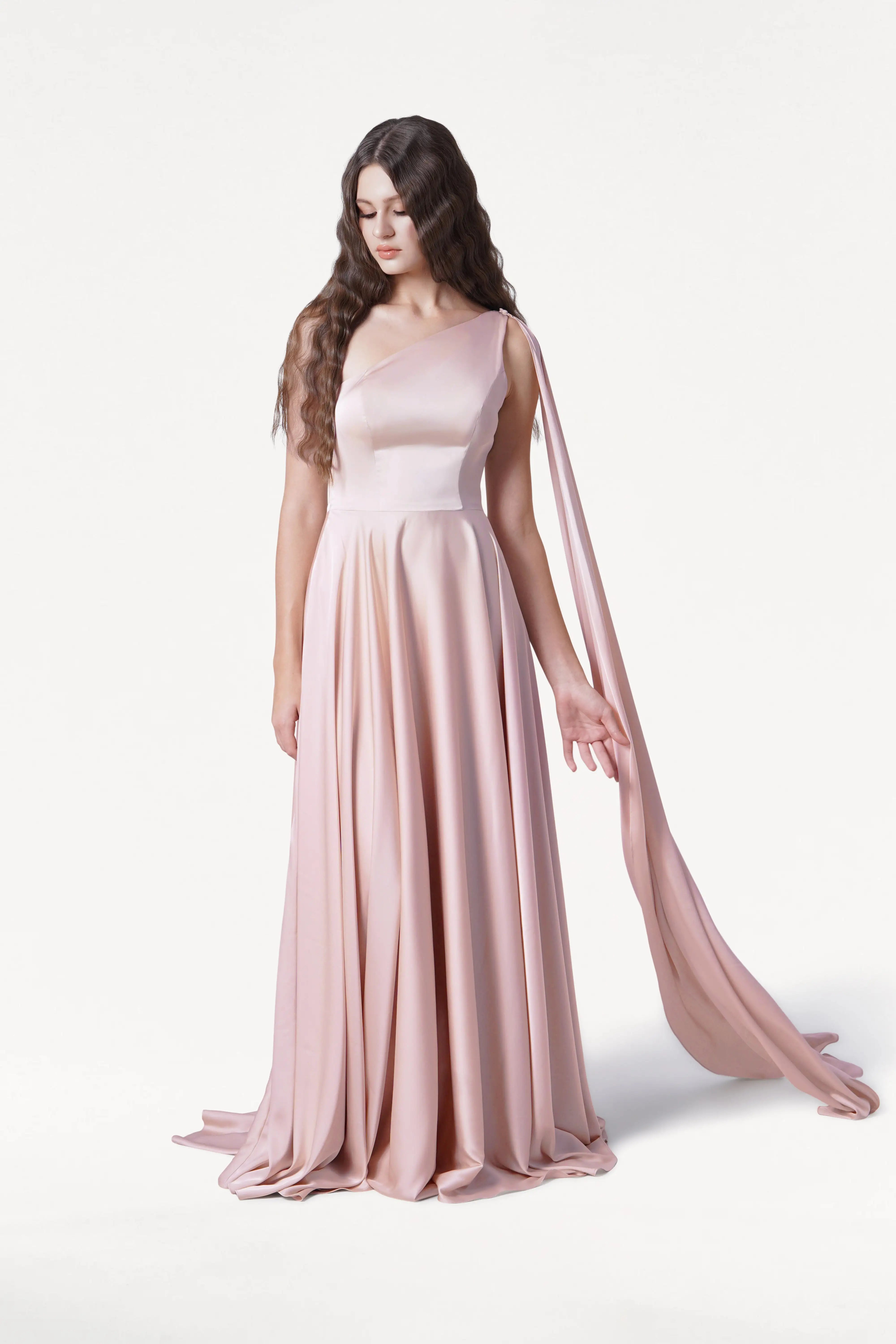 A woman with long, wavy brown hair models the TH&TH Lyra Bridesmaid Dress in blush pink satin, a flowing, size-inclusive one-shoulder style with draped sash, standing against a white background and looking down with one hand extended.