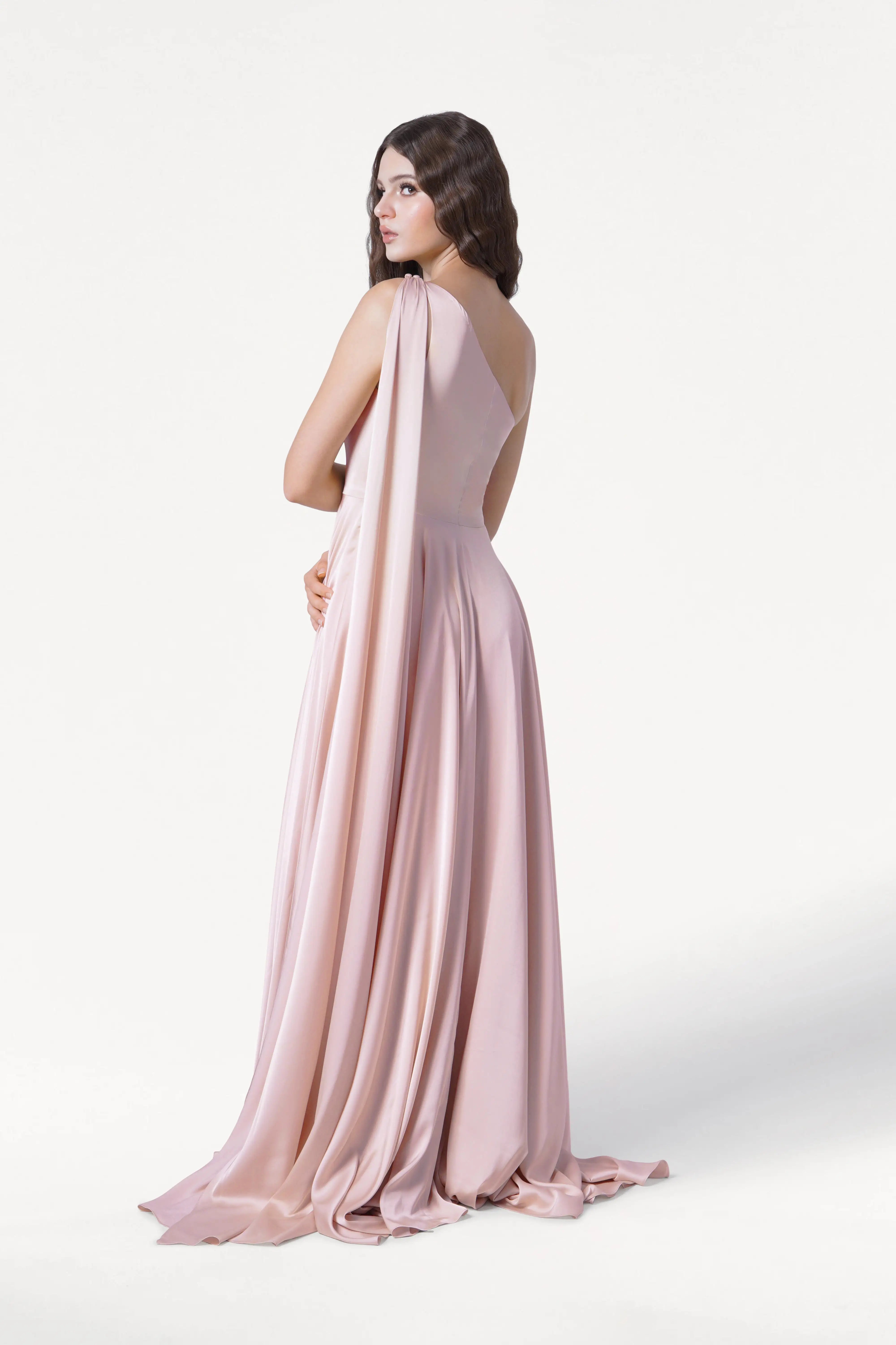 A woman models the TH&TH Lyra Bridesmaid Dress in blush pink satin, standing against a white background and looking over her shoulder with her hands gently clasped in front.