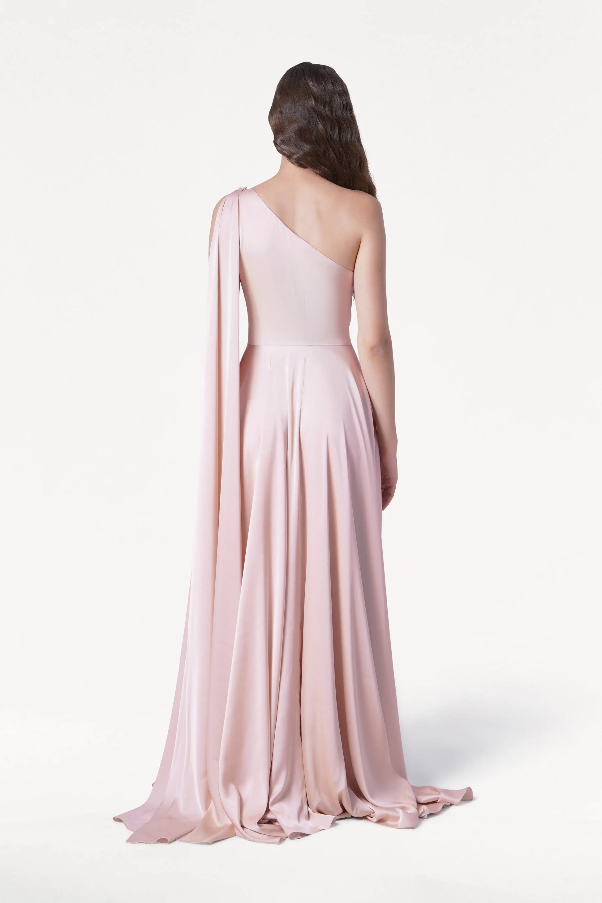 A woman with wavy brown hair stands facing away in a studio, wearing the TH&TH Lyra Bridesmaid Dress in blush pink satin—a floor-length, one-shoulder gown with a long draped sleeve.