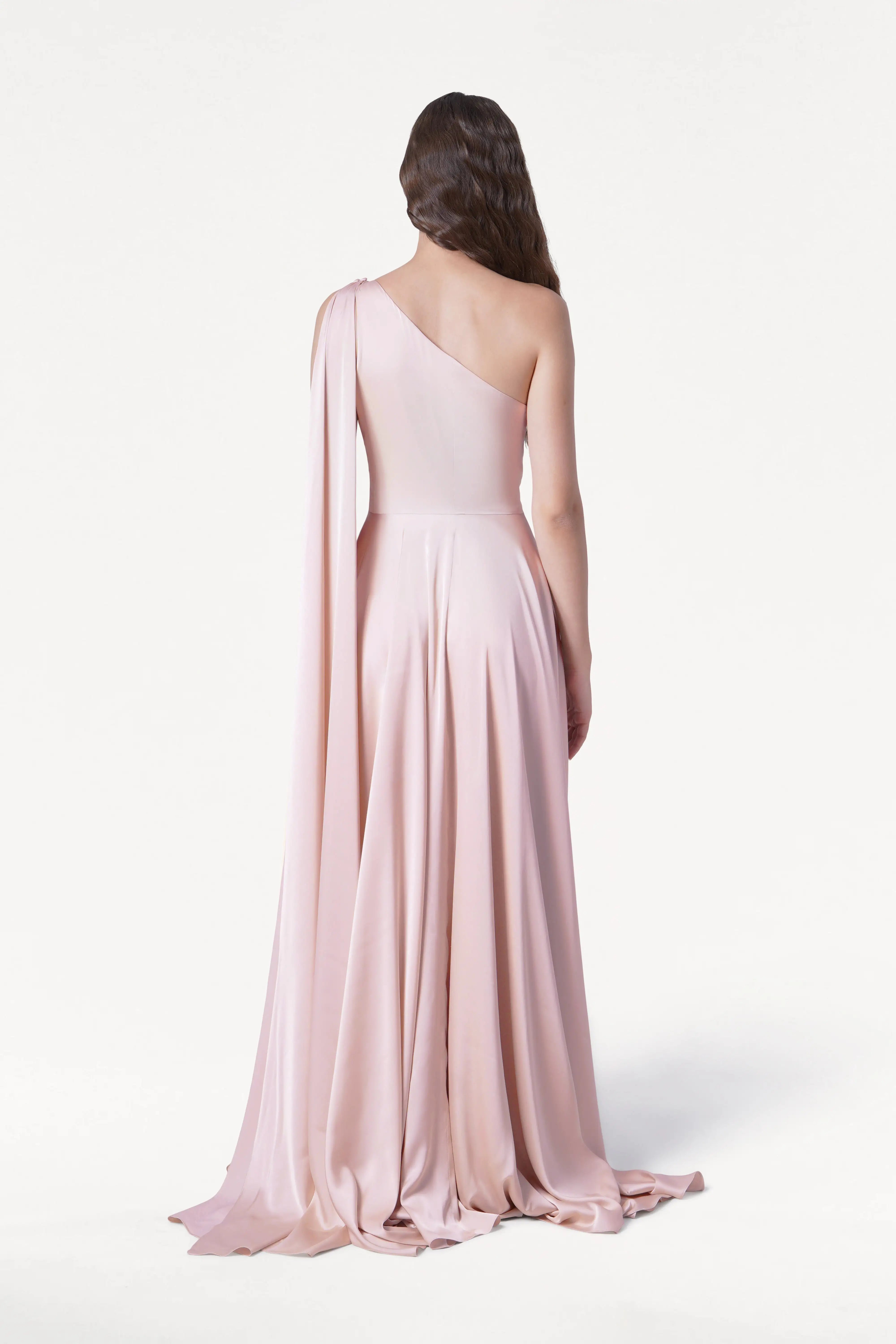A woman with wavy brown hair stands facing away in a studio, wearing the TH&TH Lyra Bridesmaid Dress in blush pink satin—a floor-length, one-shoulder gown with a long draped sleeve.
