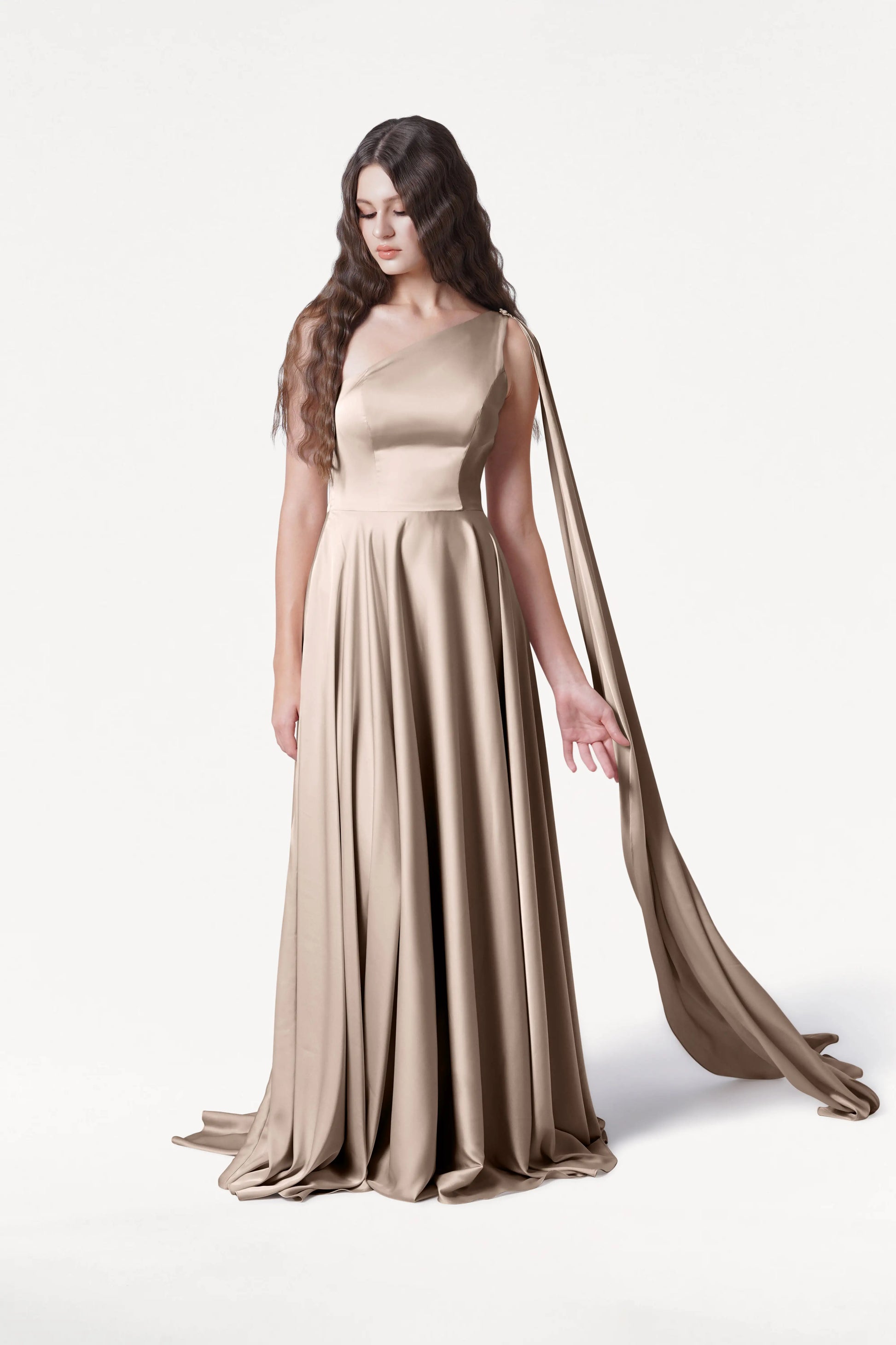 A woman with long wavy hair wears the TH&TH Lyra Bridesmaid Dress in Champagne Sky Satin, featuring a floor-length one-shoulder design and a flowing sash. She stands against a plain white background, looking down with one hand extended.