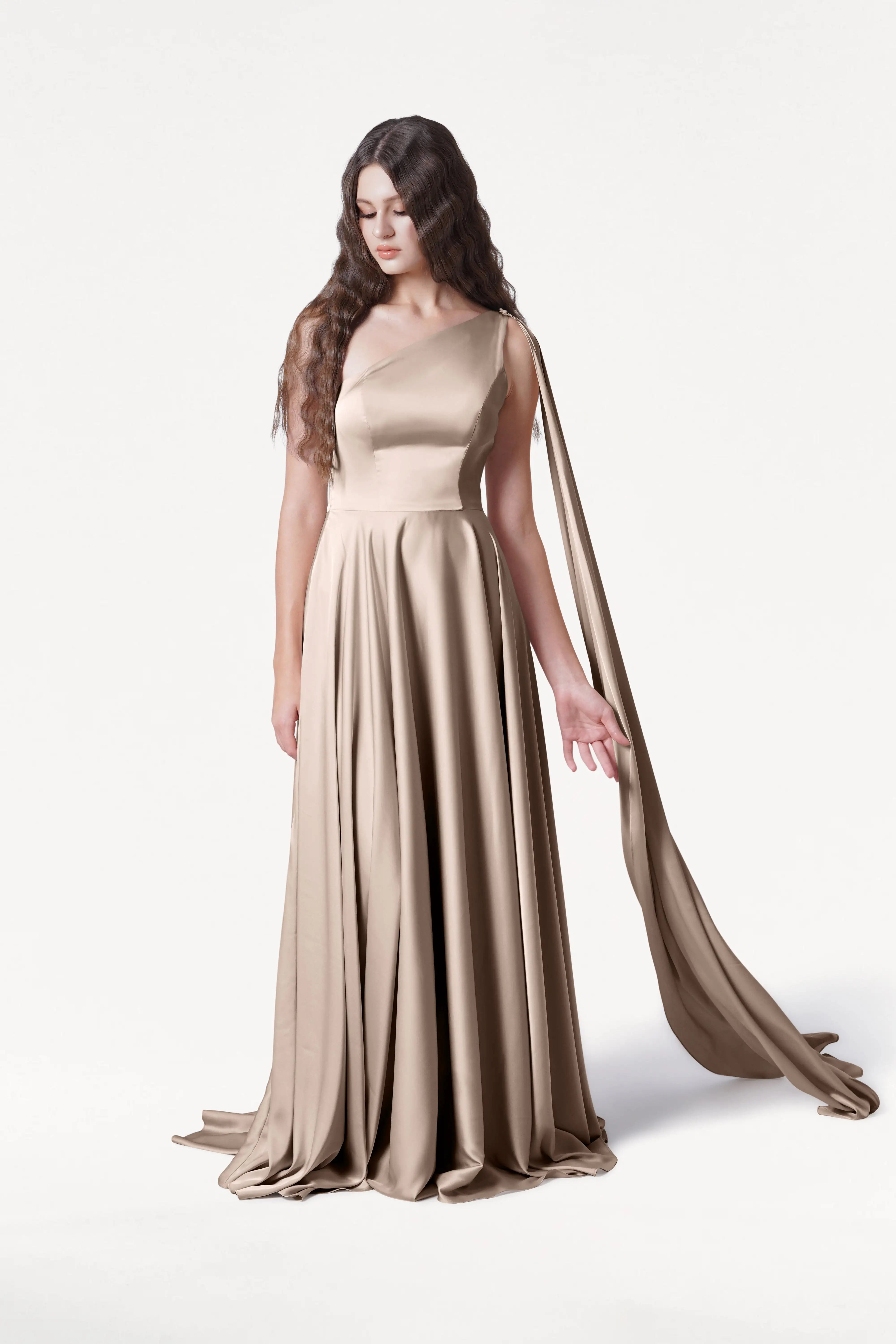 A woman with long wavy hair wears the TH&TH Lyra Bridesmaid Dress in Champagne Sky Satin, featuring a floor-length one-shoulder design and a flowing sash. She stands against a plain white background, looking down with one hand extended.