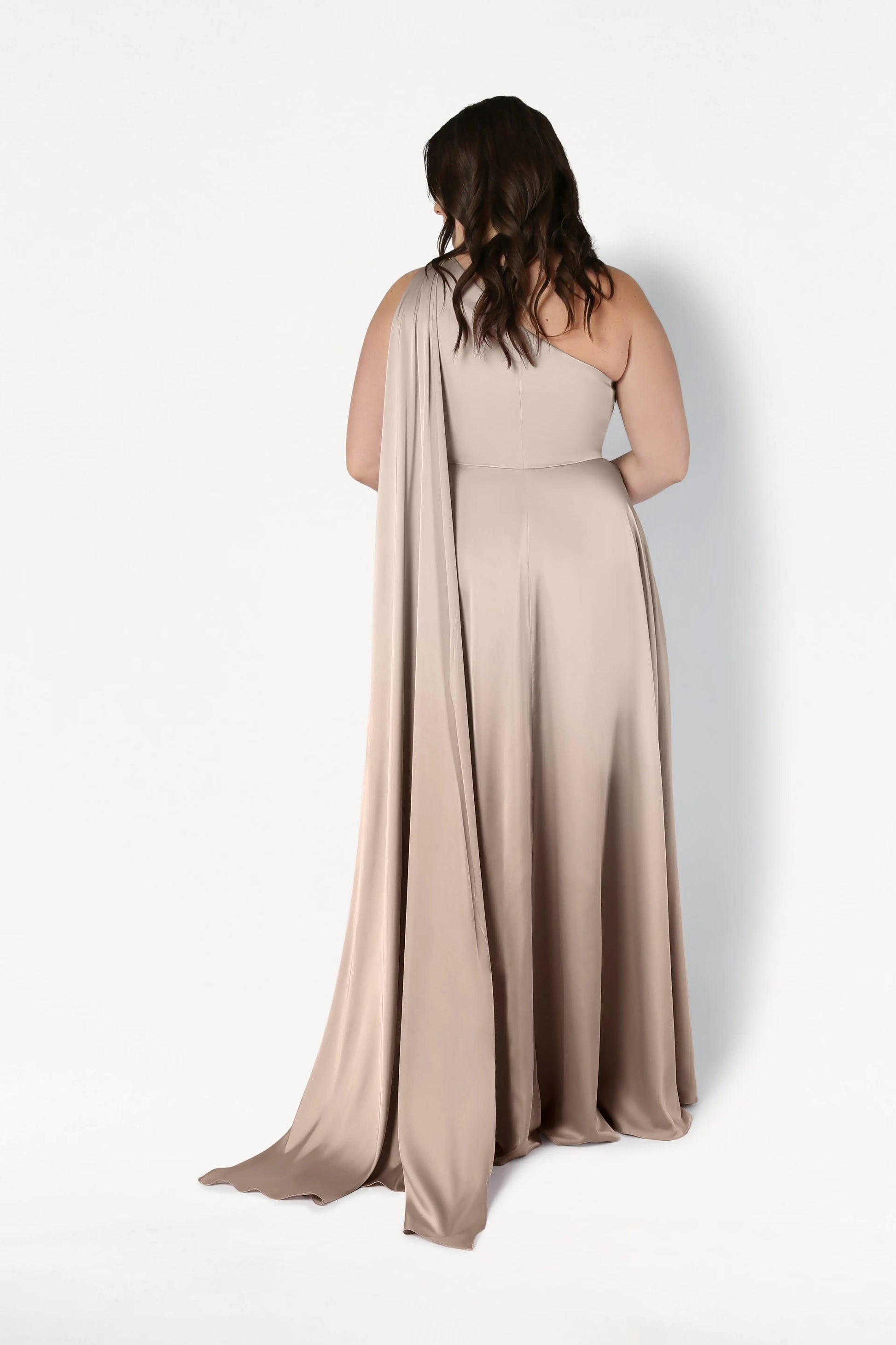 A woman with wavy brown hair stands facing a white wall, her back to the camera, wearing the TH&TH Lyra Bridesmaid Dress in Champagne Sky Satin—a size-inclusive, flowing one-shoulder gown with a dramatic draped train.