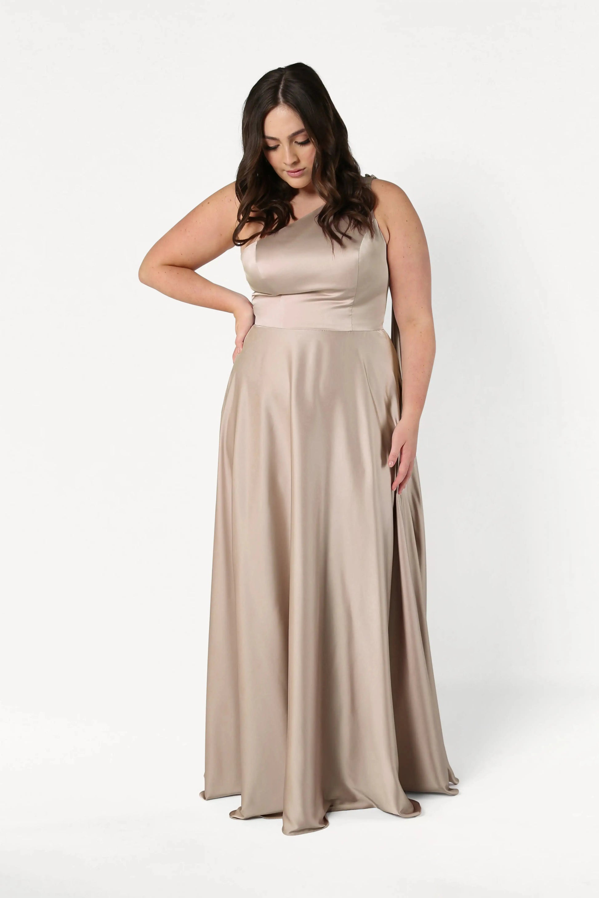 A woman with long dark hair models the TH&TH Lyra Bridesmaid Dress in Champagne Sky Satin, a sleeveless, floor-length gown, standing against a plain white background and looking down with one hand on her hip.