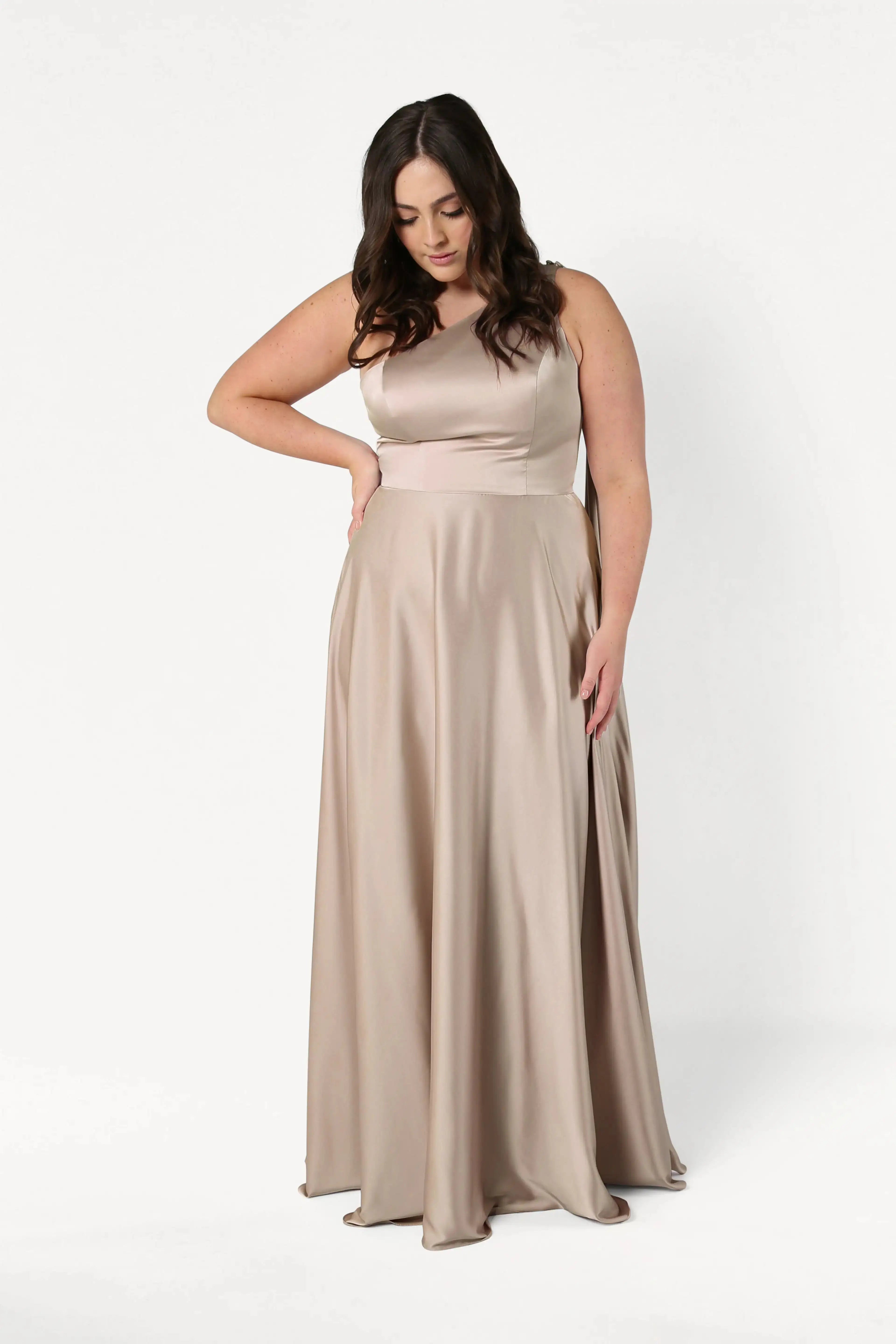 A woman with long dark hair models the TH&TH Lyra Bridesmaid Dress in Champagne Sky Satin, a sleeveless, floor-length gown, standing against a plain white background and looking down with one hand on her hip.