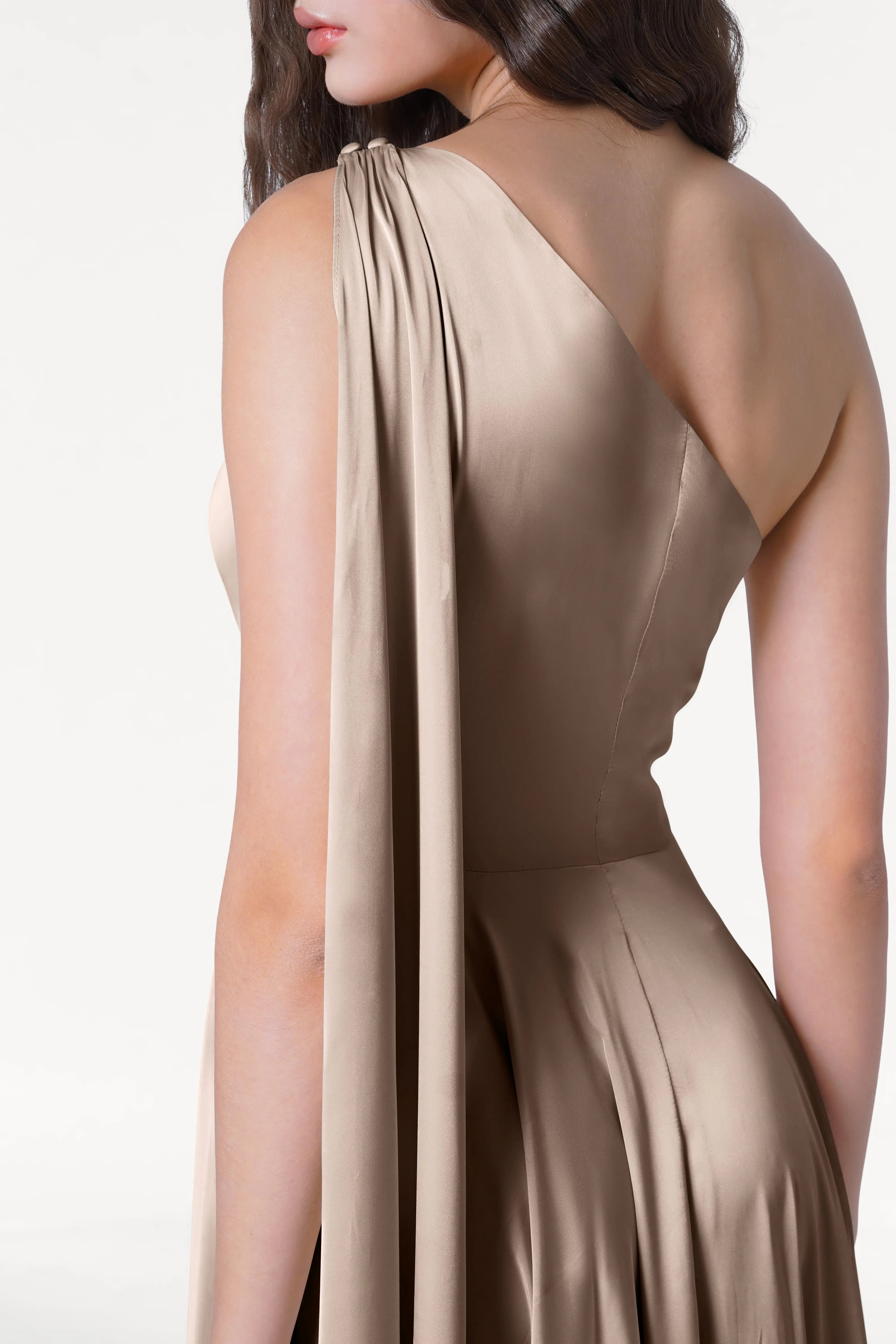 A woman models the Lyra Bridesmaid Dress in Champagne Sky Satin by TH&TH, featuring a shiny beige finish and draped one-shoulder sleeve, against a plain white background. Her long wavy hair cascades down, partly obscuring her face.