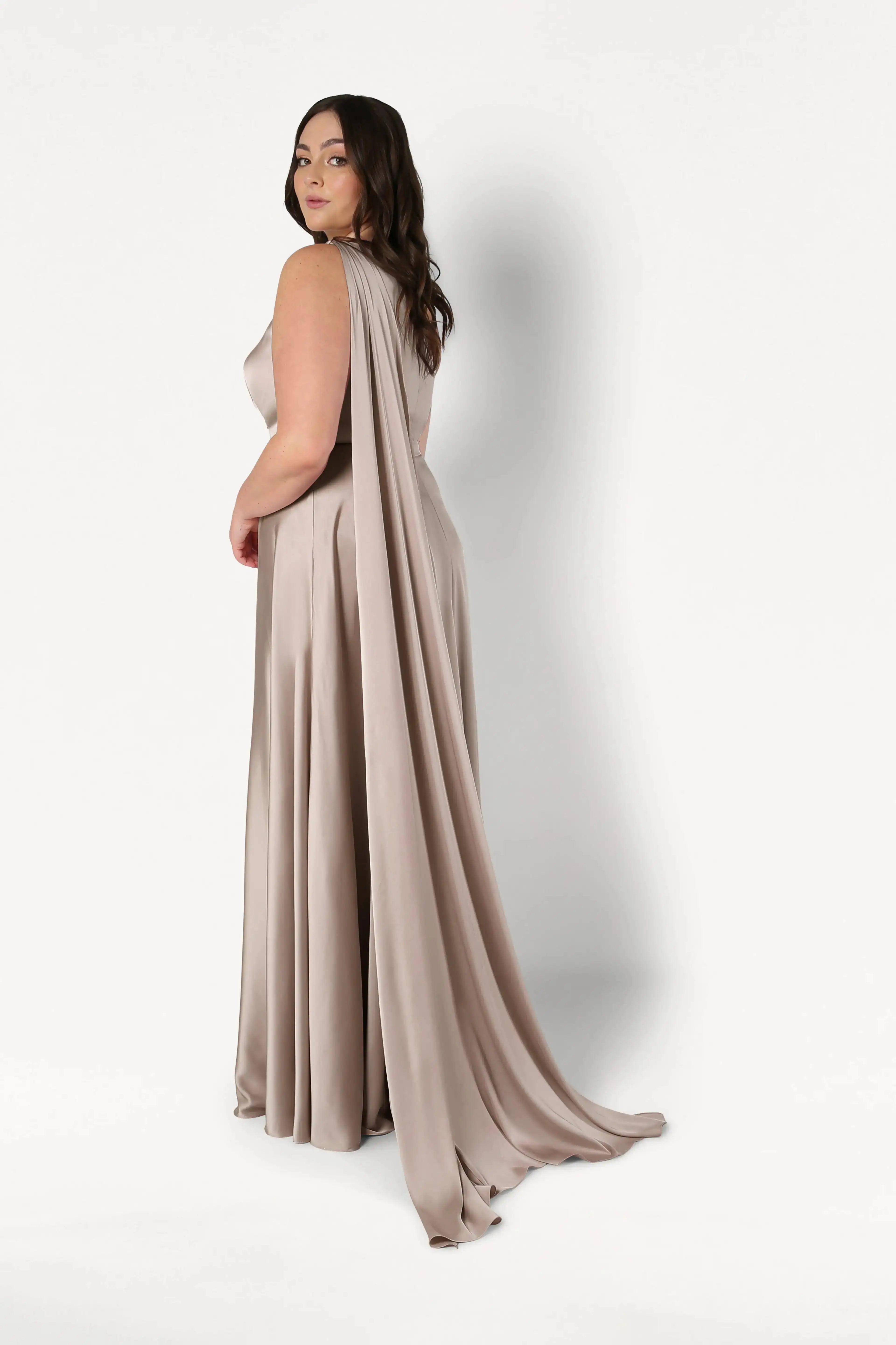 A woman wears the Lyra Bridesmaid Dress in Champagne Sky Satin by TH&TH— a taupe, floor-length gown with a long draped cape— and stands against a white background, glancing over her shoulder.