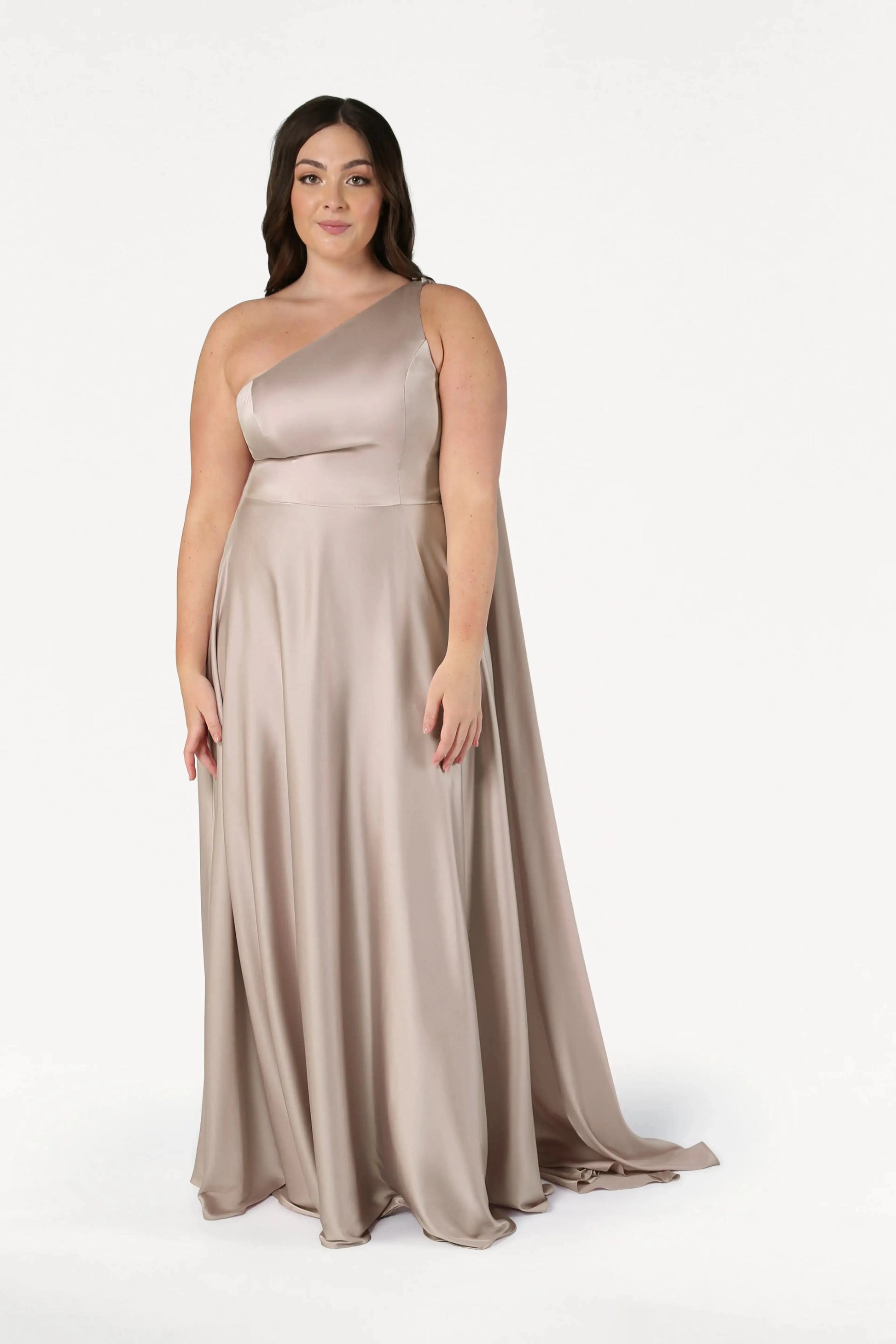 A woman with long dark hair wears the TH&TH Lyra Bridesmaid Dress in Champagne Sky Satin—a size-inclusive, one-shoulder gown. She stands facing forward against a plain white background, arms relaxed by her sides.
