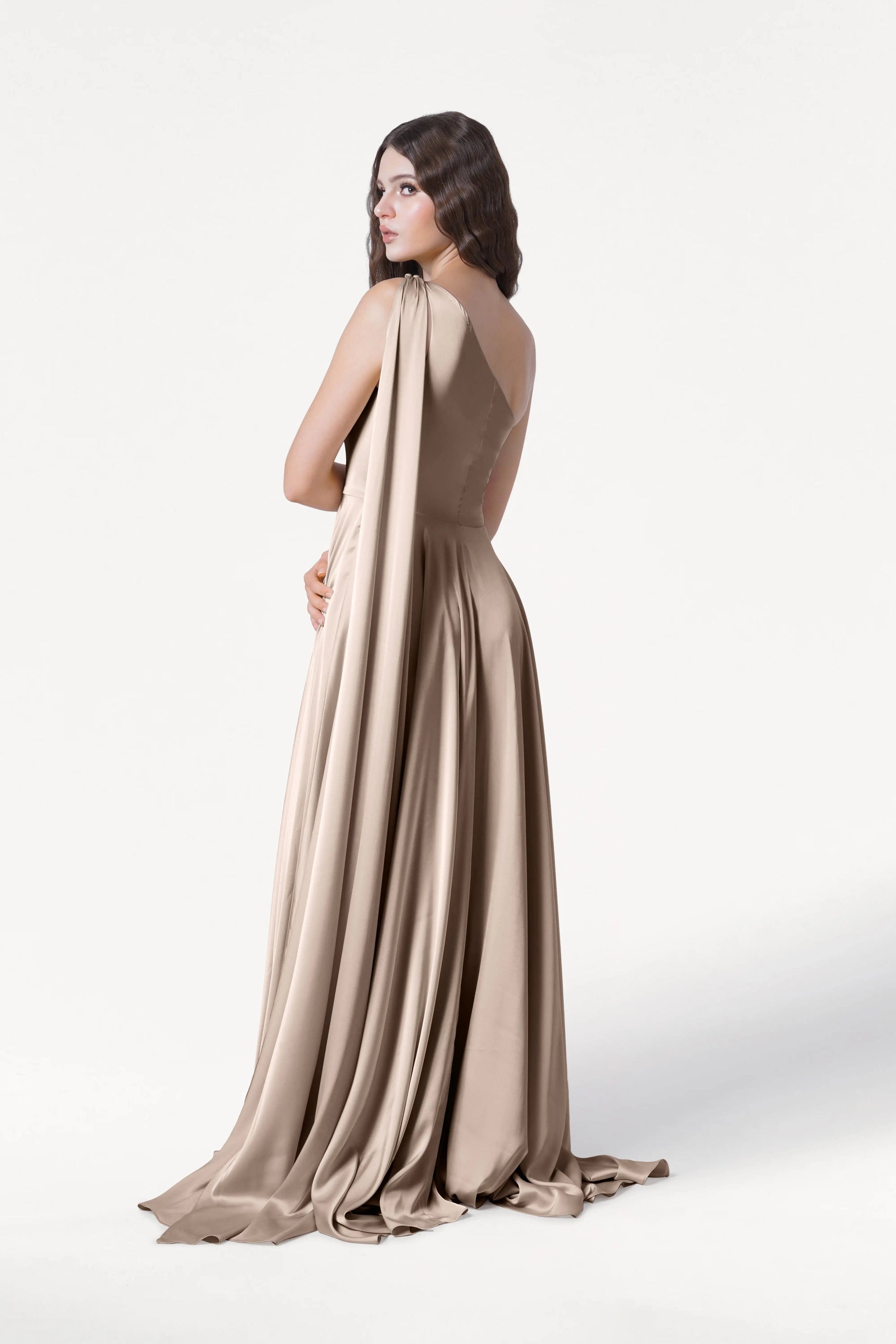 A woman models the TH&TH Lyra Bridesmaid Dress in Champagne Sky Satin, a size-inclusive gown with a flowing train and draped satin fabric, standing with her back to the camera against a plain white background.