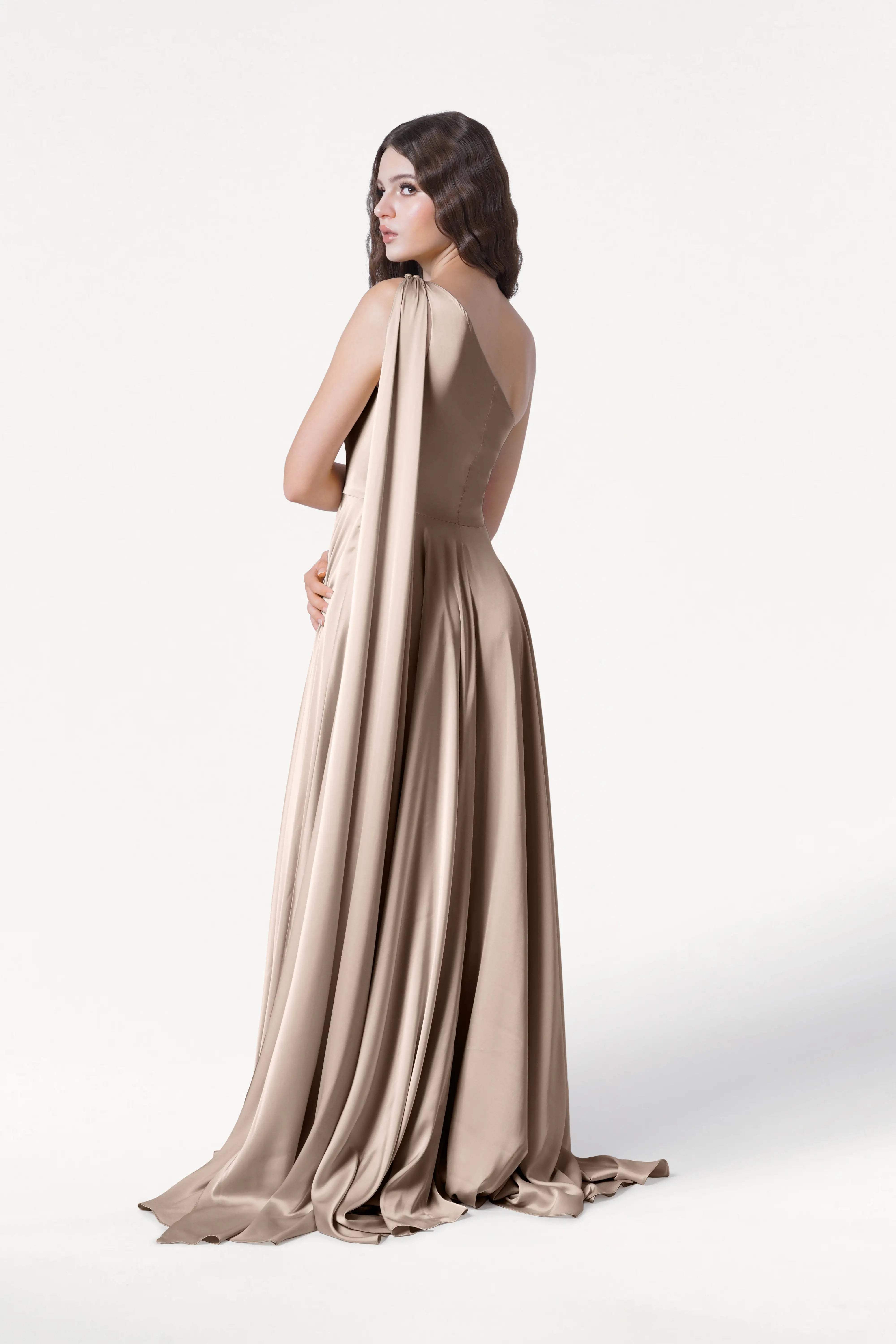 A woman models the TH&TH Lyra Bridesmaid Dress in Champagne Sky Satin, a size-inclusive gown with a flowing train and draped satin fabric, standing with her back to the camera against a plain white background.
