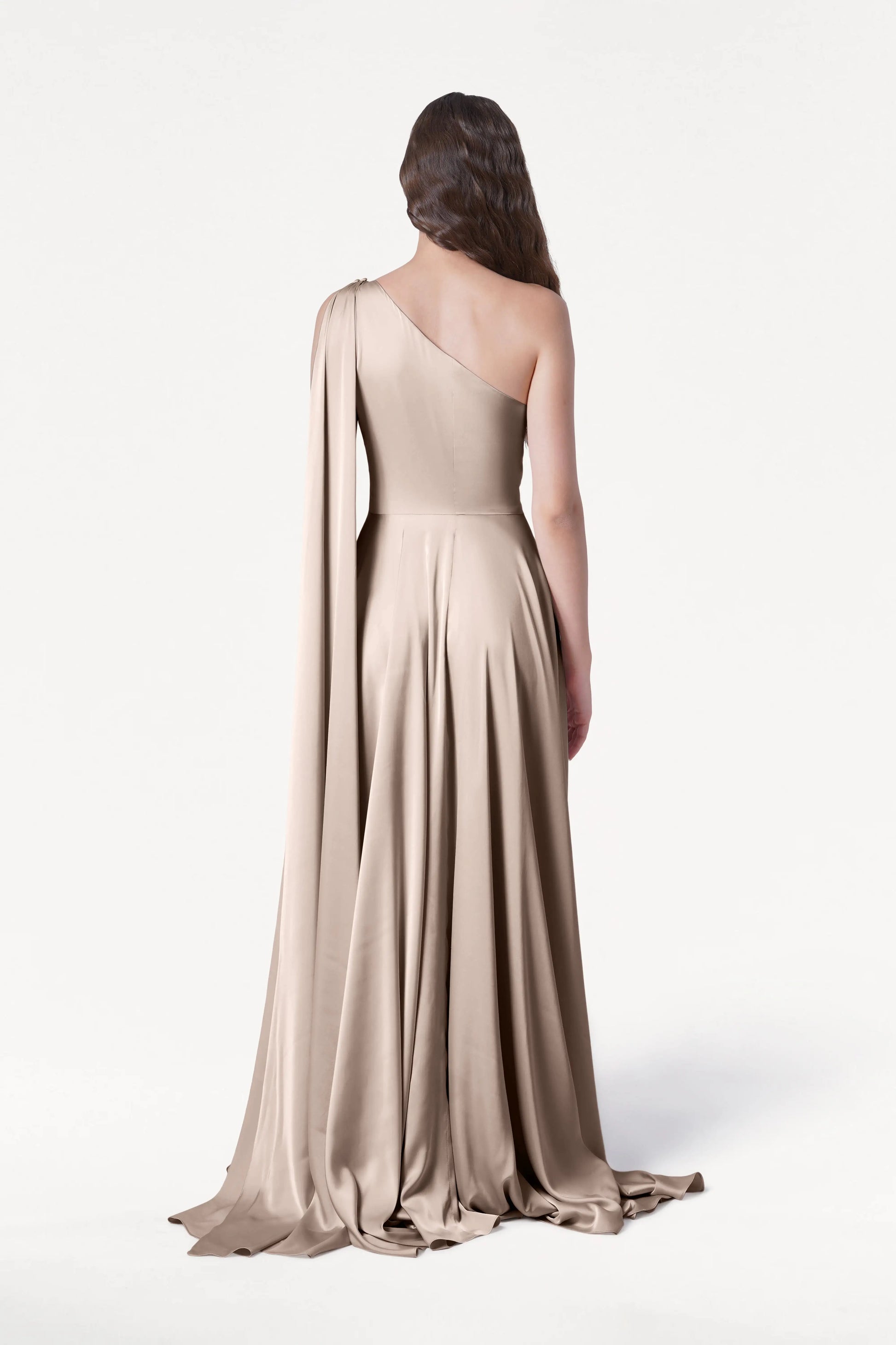 A woman faces away, wearing the TH&TH Lyra Bridesmaid Dress in Champagne Sky Satin—a floor-length, one-shoulder gown with a draped sash—set against a plain white background.