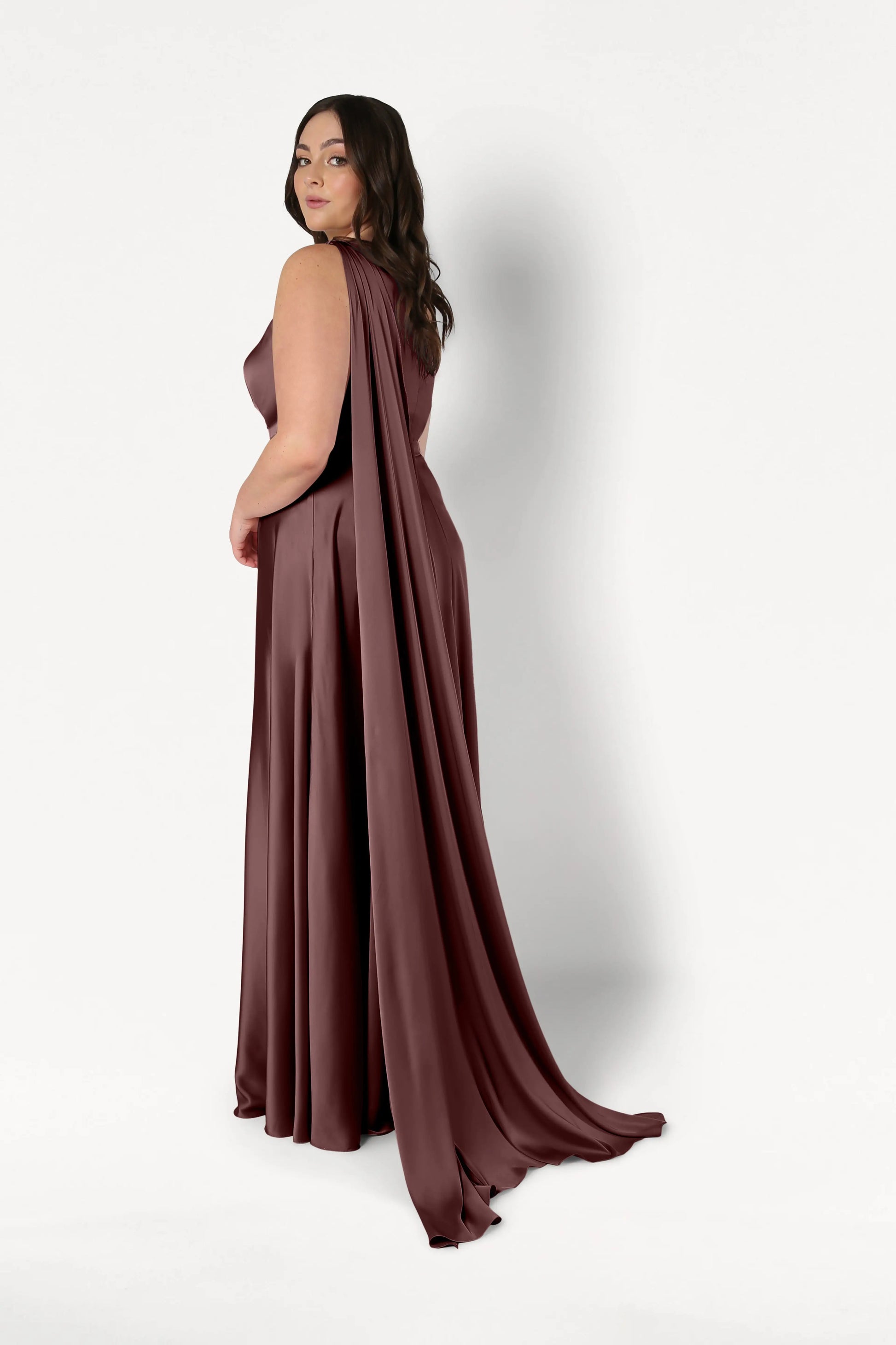 A woman in the TH&TH Lyra Bridesmaid Dress, a size-inclusive, floor-length chocolate brown satin gown with a long cape, stands against a plain white background, looking over her shoulder.