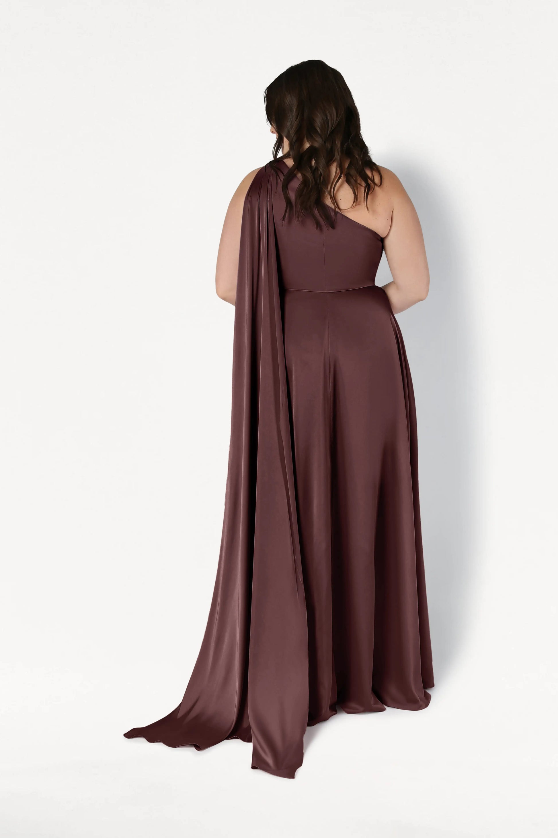 A woman with wavy brown hair models the TH&TH Lyra Bridesmaid Dress in chocolate brown satin. The size-inclusive, floor-length one-shoulder gown features a long, draped sash, shown from the back against a white background.