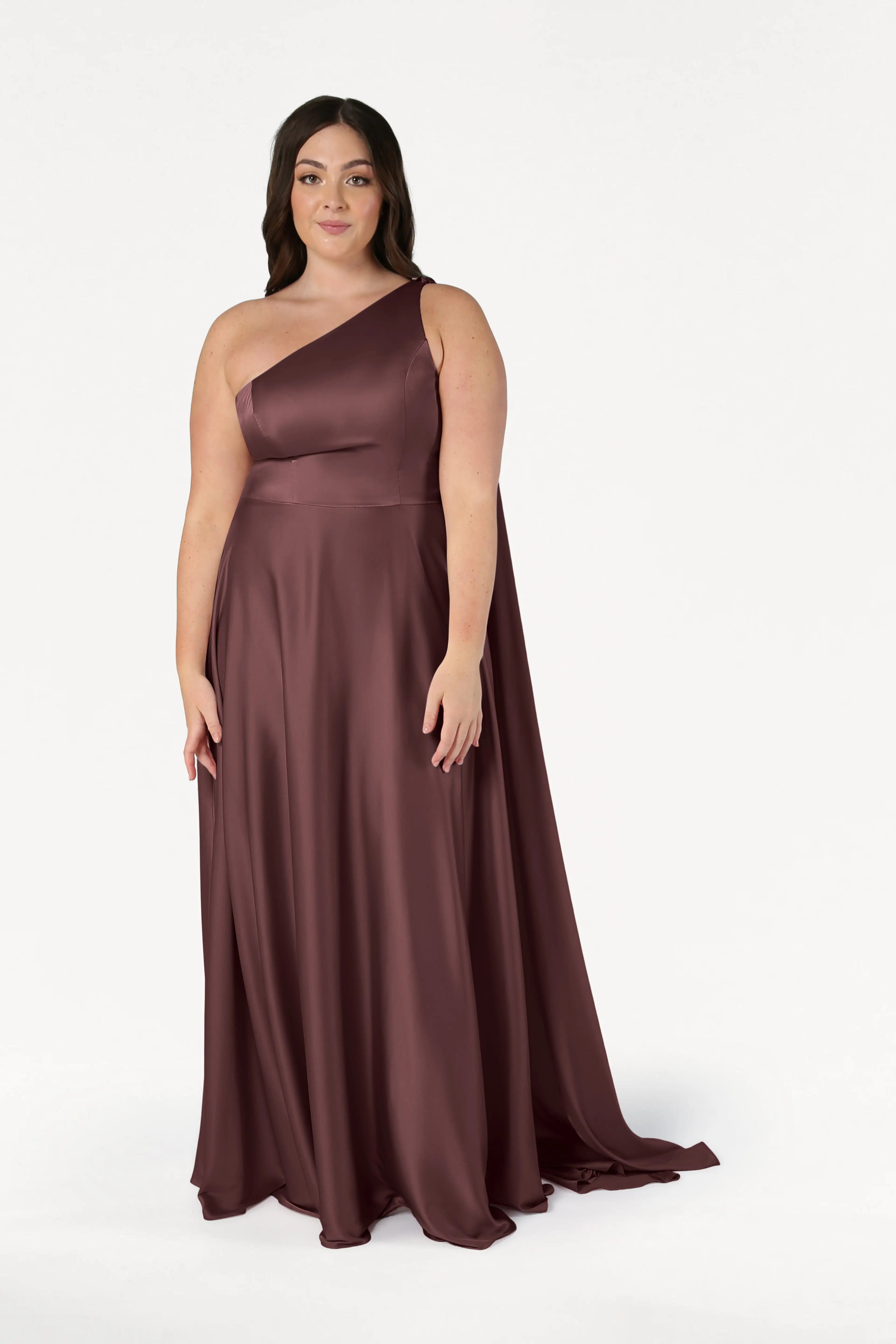 A woman wears the size-inclusive Lyra Bridesmaid Dress by TH&TH in chocolate brown satin, featuring a floor-length, one-shoulder design, as she stands against a plain white background with a slight smile.