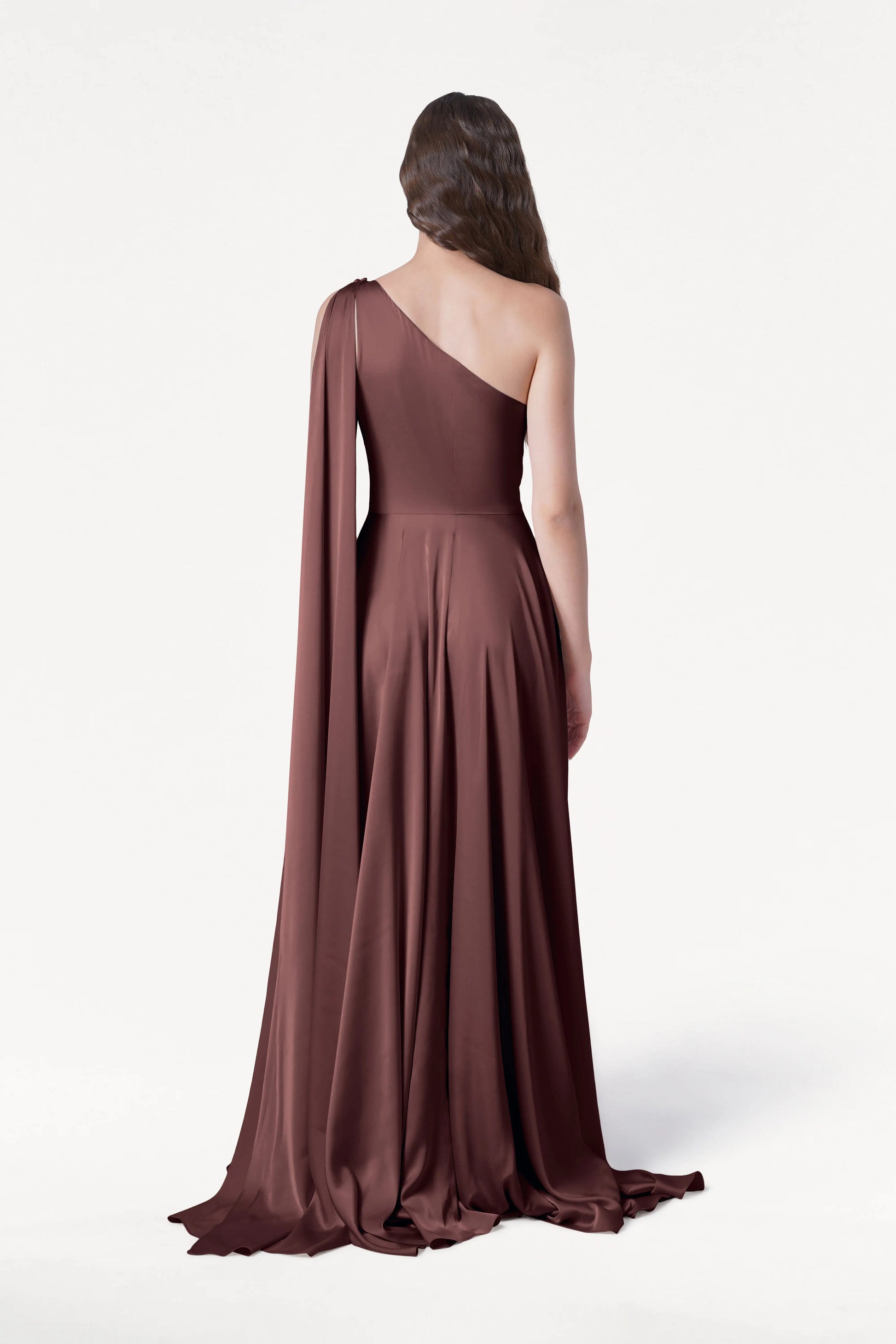 A person with long wavy hair models the Lyra Bridesmaid Dress in Chocolate Brown Satin by TH&TH—a size-inclusive, floor-length one-shoulder gown with a draped cape—facing away from the camera against a plain white background.
