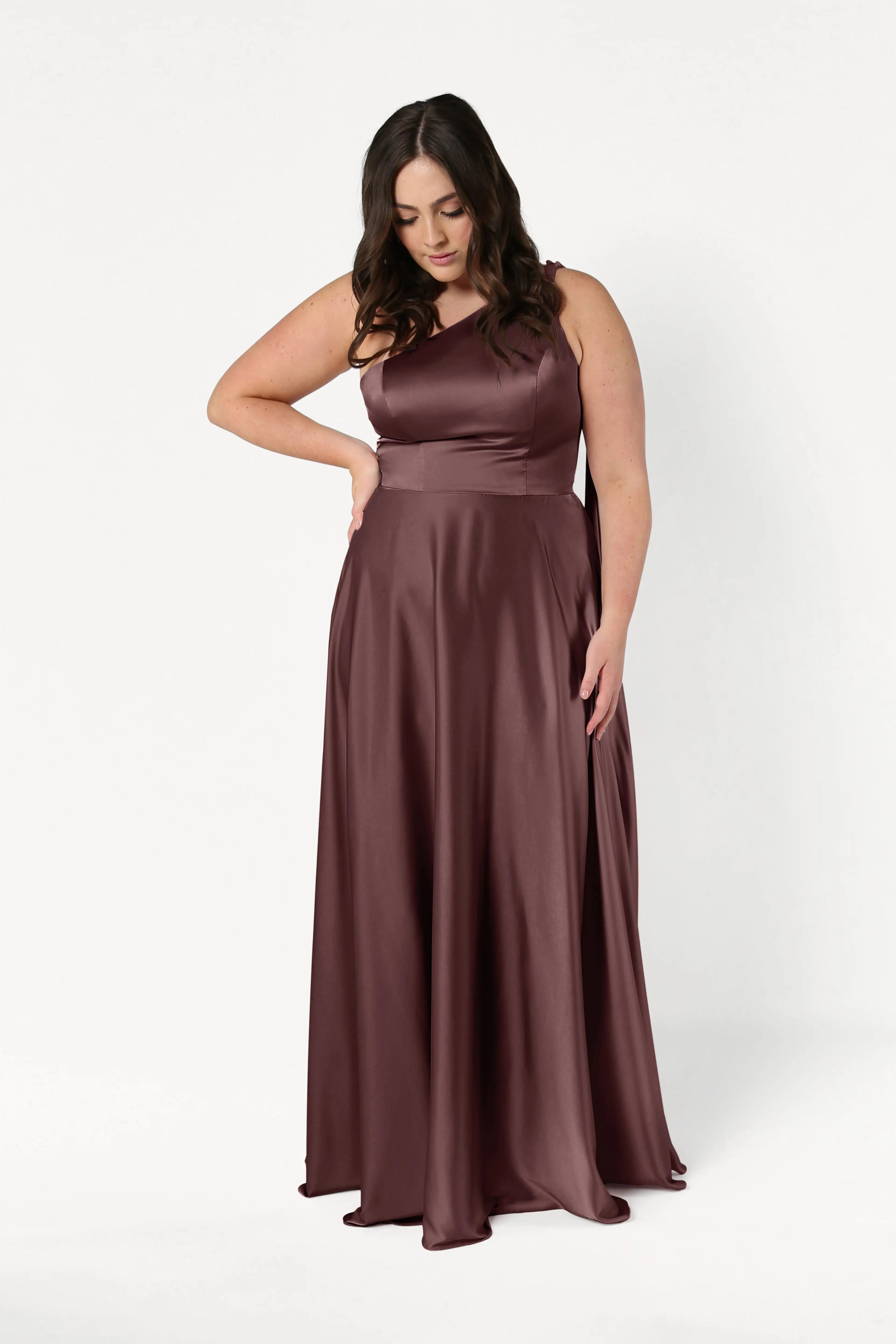 A woman wears the TH&TH Lyra Bridesmaid Dress in chocolate brown satin, a size-inclusive sleeveless floor-length gown. She stands against a white background, looking down with one hand on her waist and loose waves in her hair.
