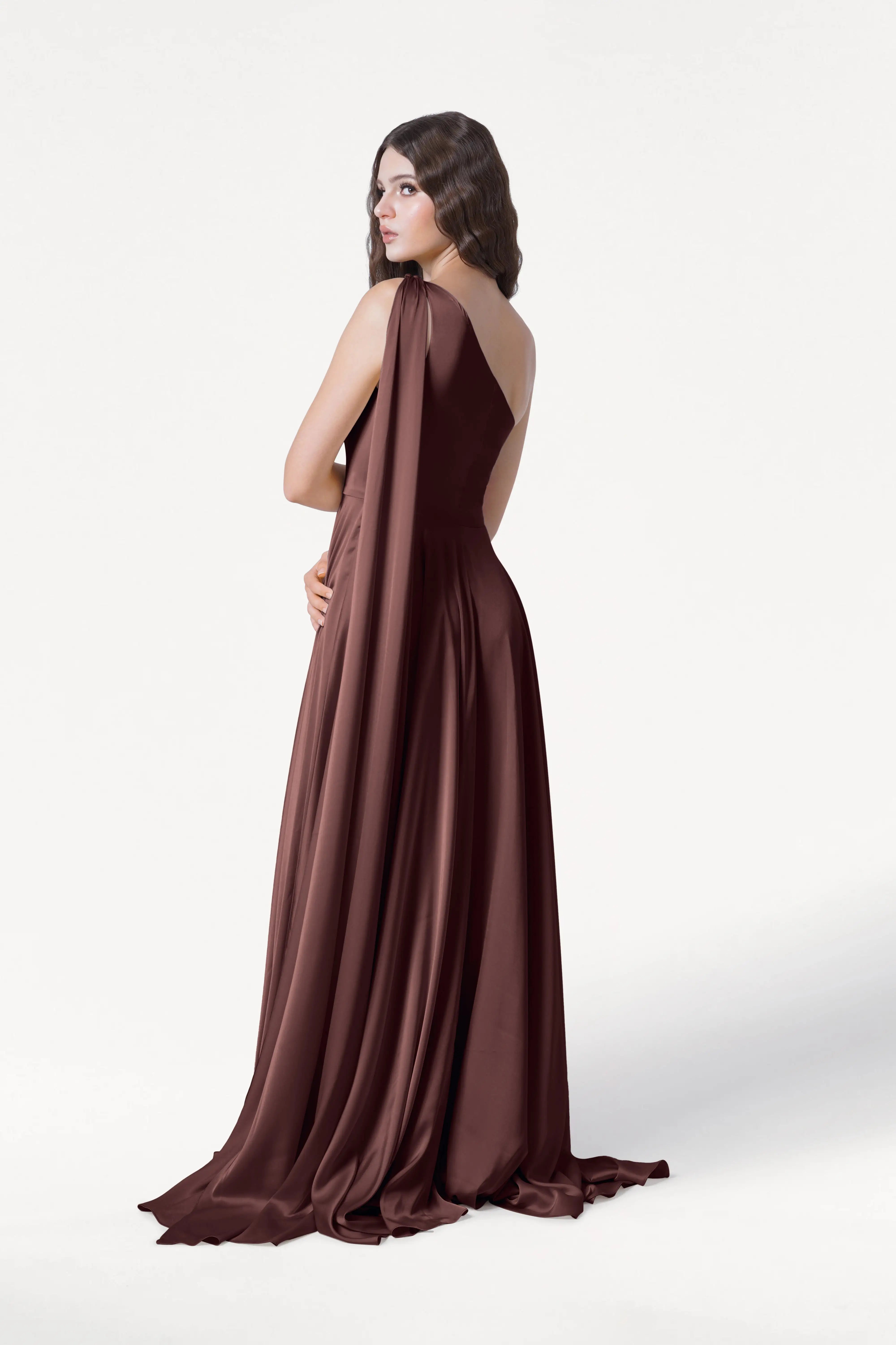 A woman models the TH&TH Lyra Bridesmaid Dress in Chocolate Brown Satin, standing with her back turned and head tilted toward the camera against a plain white background.