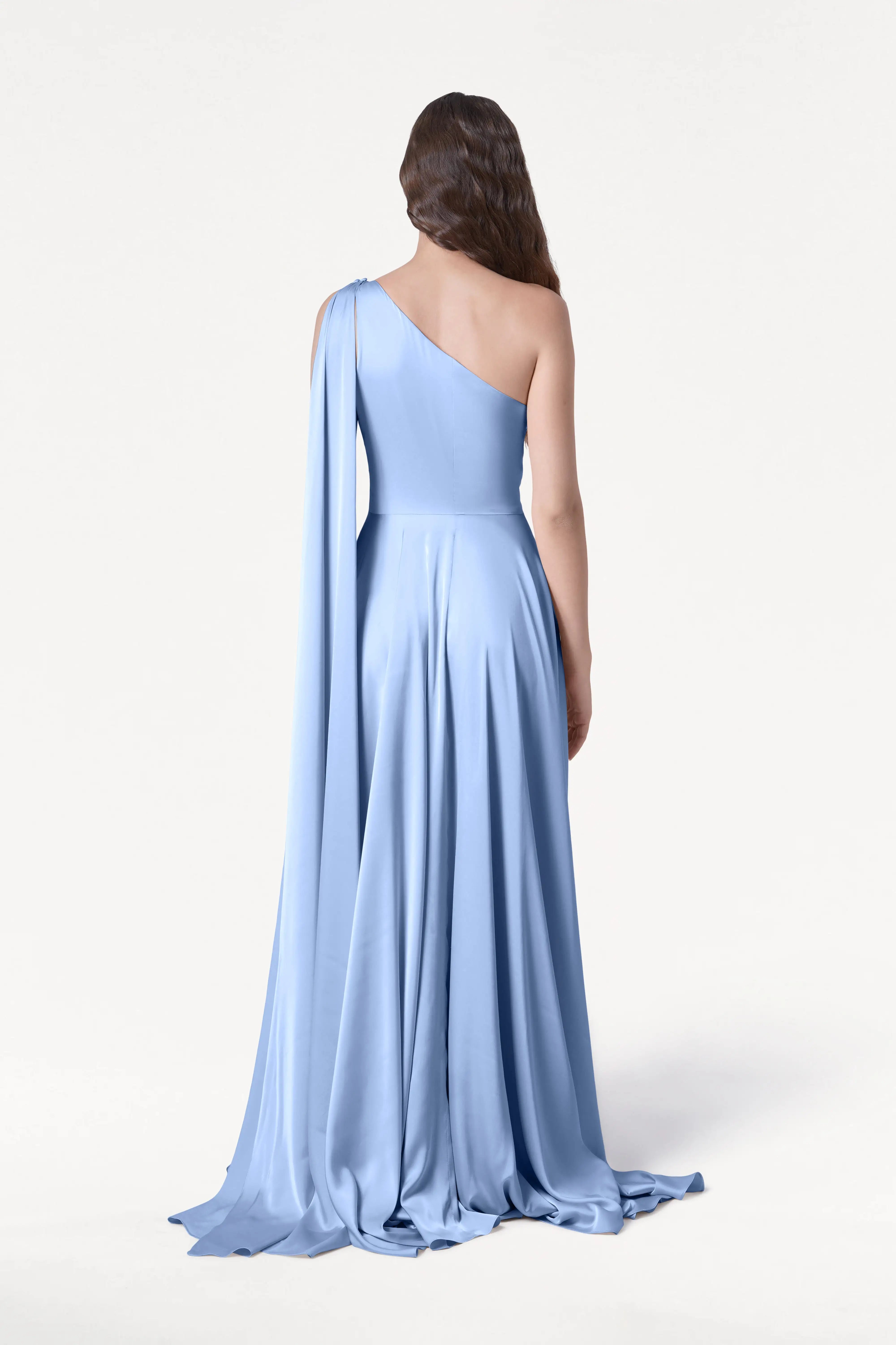 A woman with loose waves faces away in the Lyra Bridesmaid Dress by TH&TH, featuring light blue satin, one-shoulder draped sleeve, and a size-inclusive fit. The background is plain white, highlighting the gown's effortless elegance.