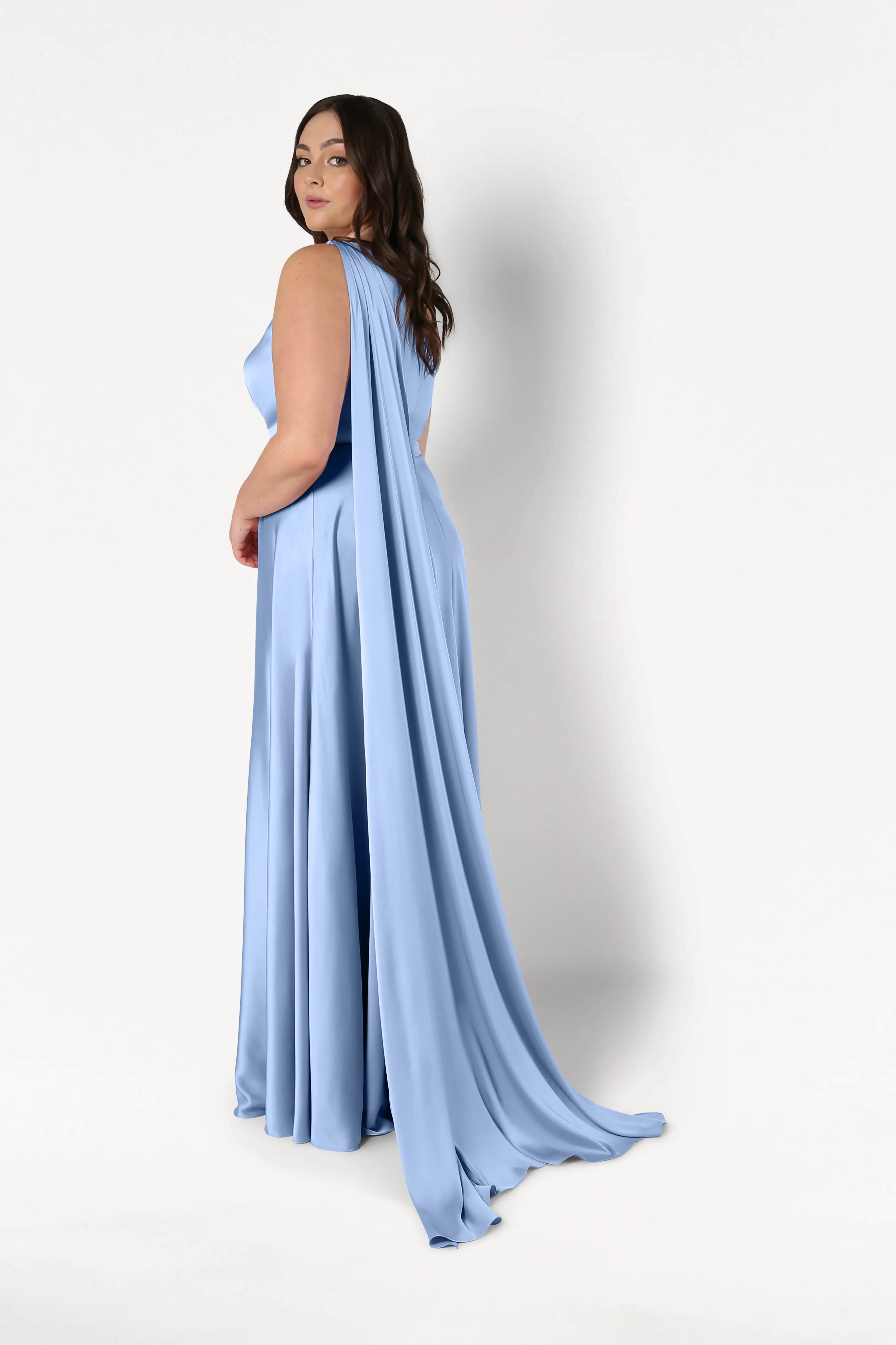 A woman models the Lyra Bridesmaid Dress in Dusty Blue Satin by TH&TH, featuring a flowing, size-inclusive design with a long draped cape, as she poses against a plain white background looking over her shoulder.